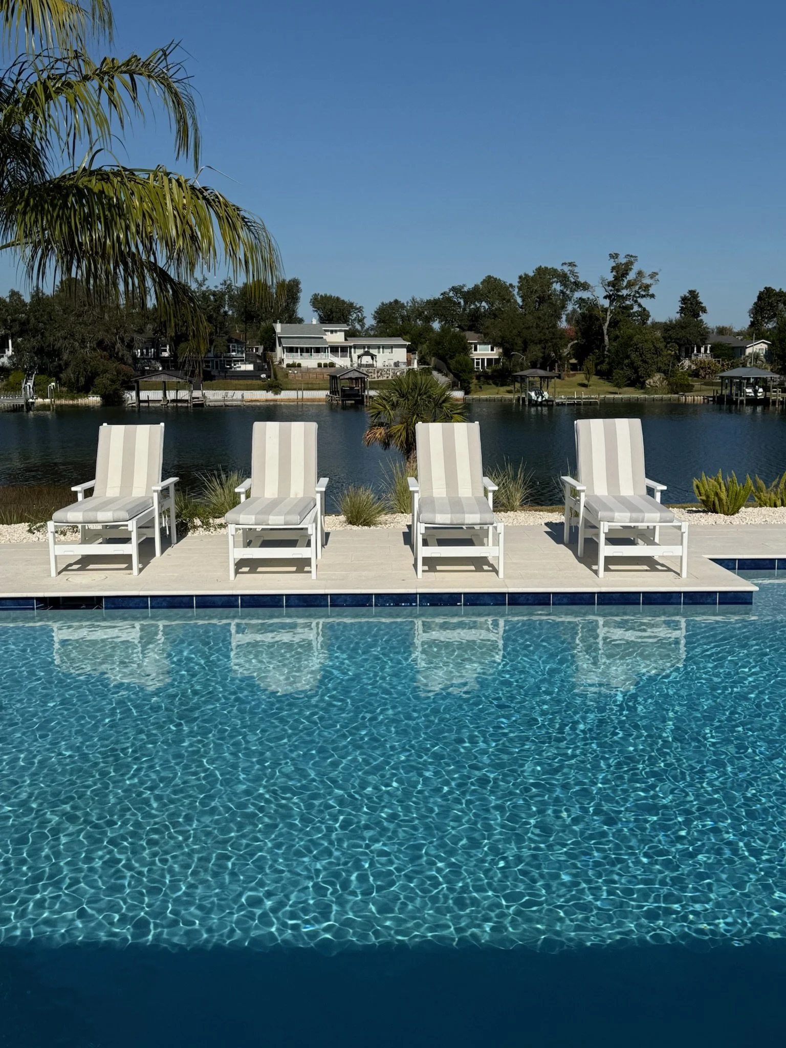 A swimming pool with clear blue water and four white lounge chairs in the foreground, overlooking a lake with houses and trees in the background under a clear blue sky.