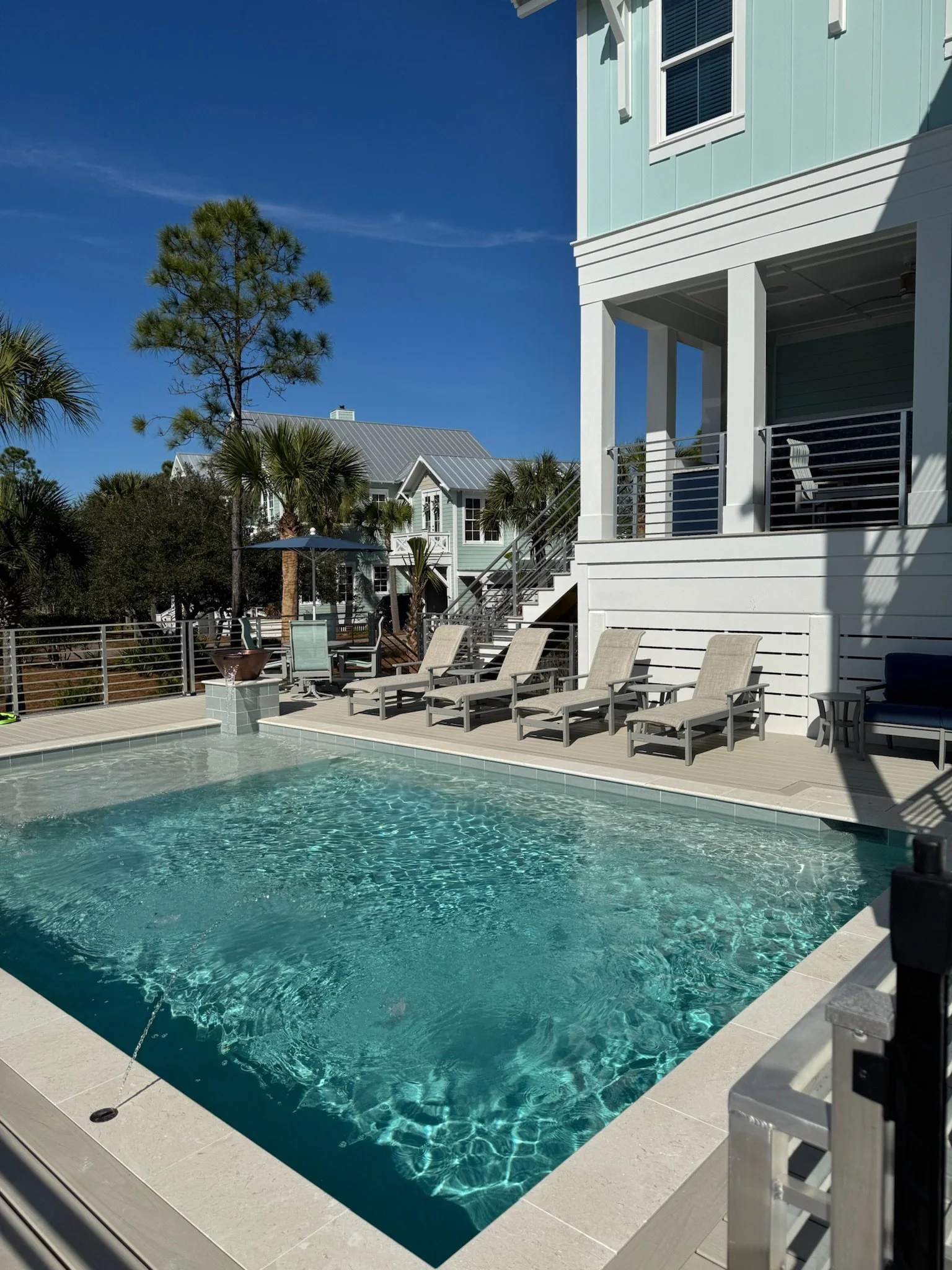 View of a backyard swimming pool with lounge chairs, surrounded by a white deck and a modern house with pastel blue siding, under a clear blue sky with some trees in the background.