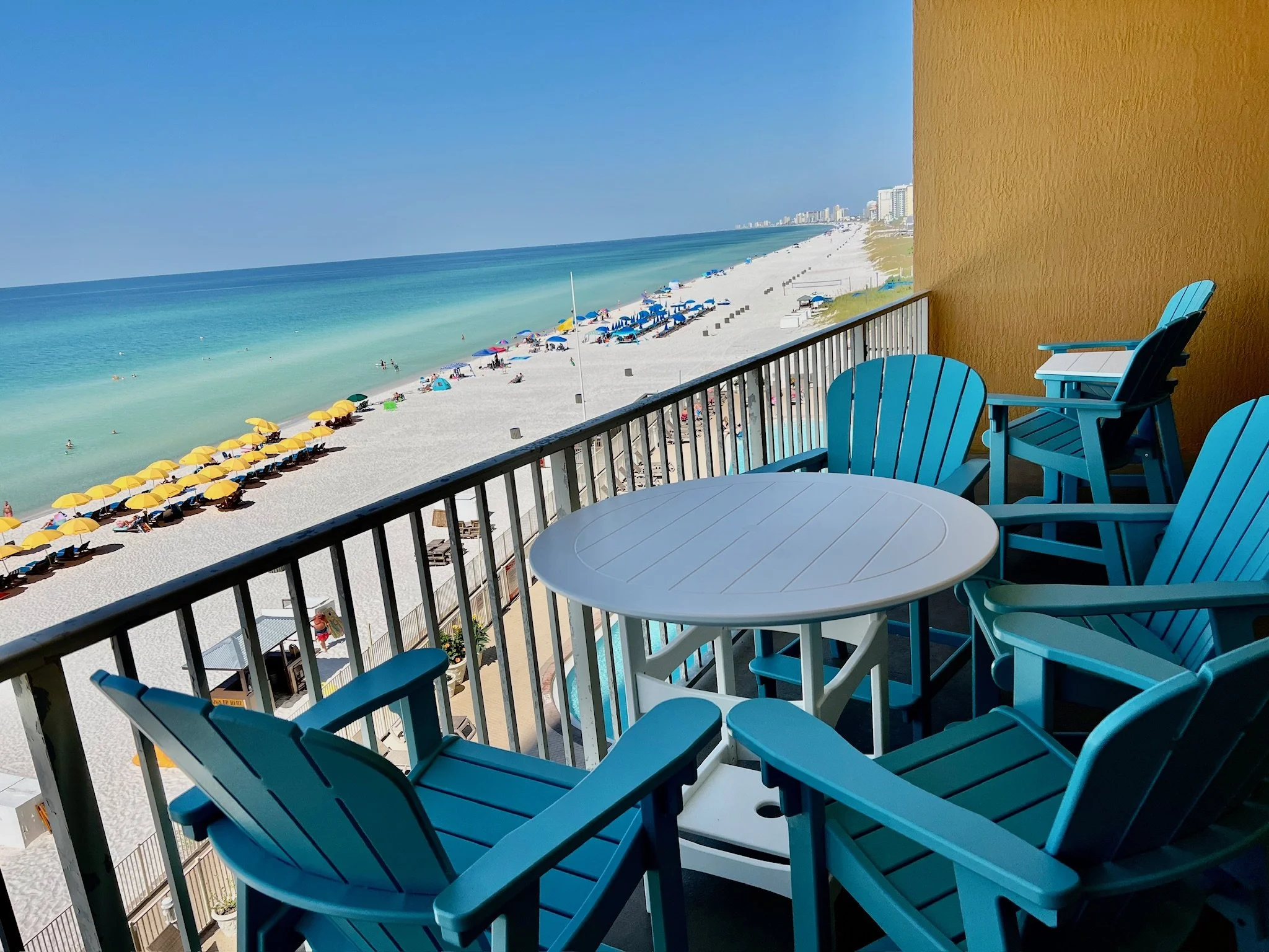 Balcony view overlooking a sandy beach with yellow and blue umbrellas, with the ocean and city skyline in the background.
