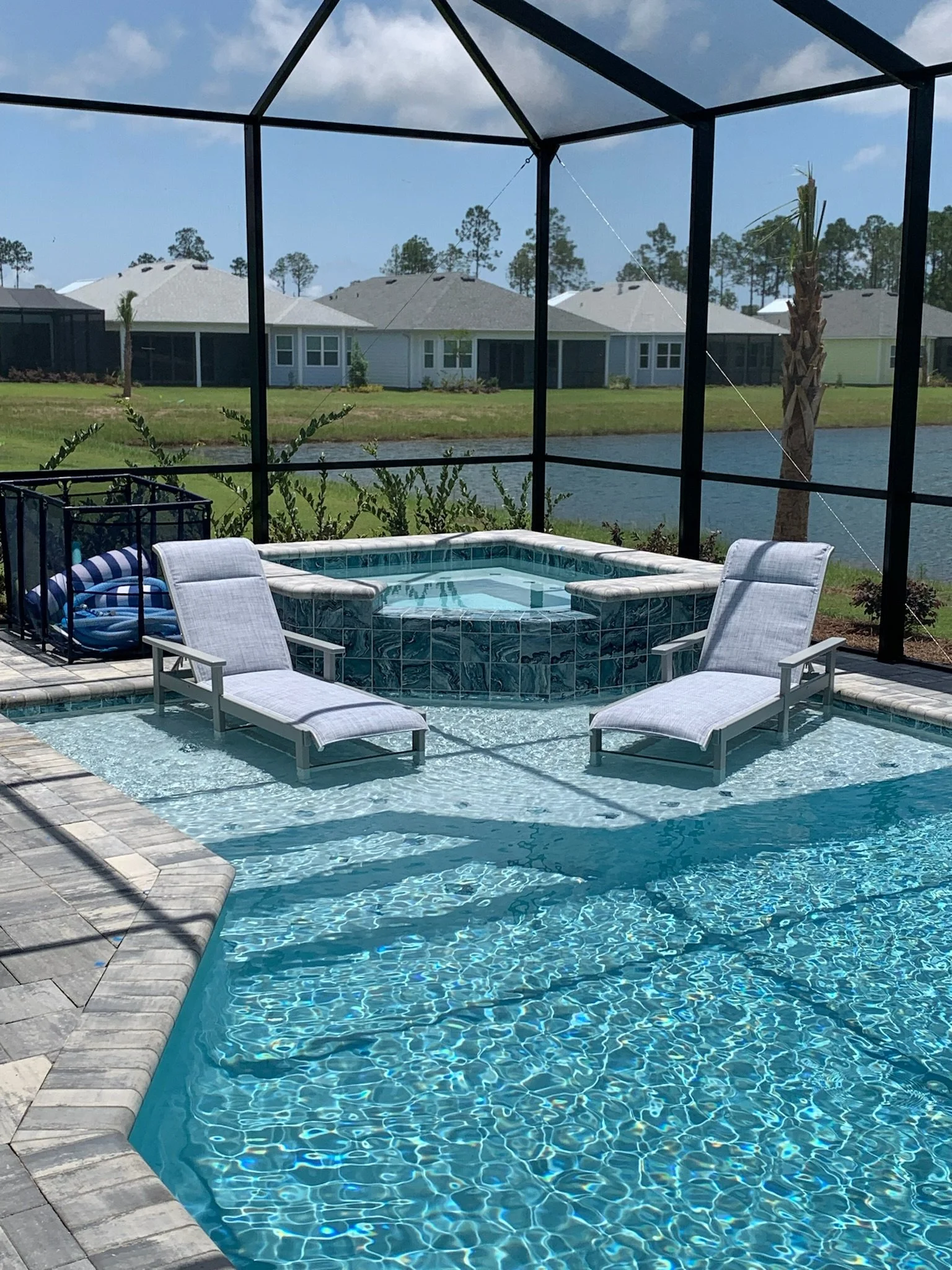 A backyard swimming pool with two lounge chairs on the pool deck, a hot tub, and a screened enclosure. In the background, there are houses, trees, and a pond.