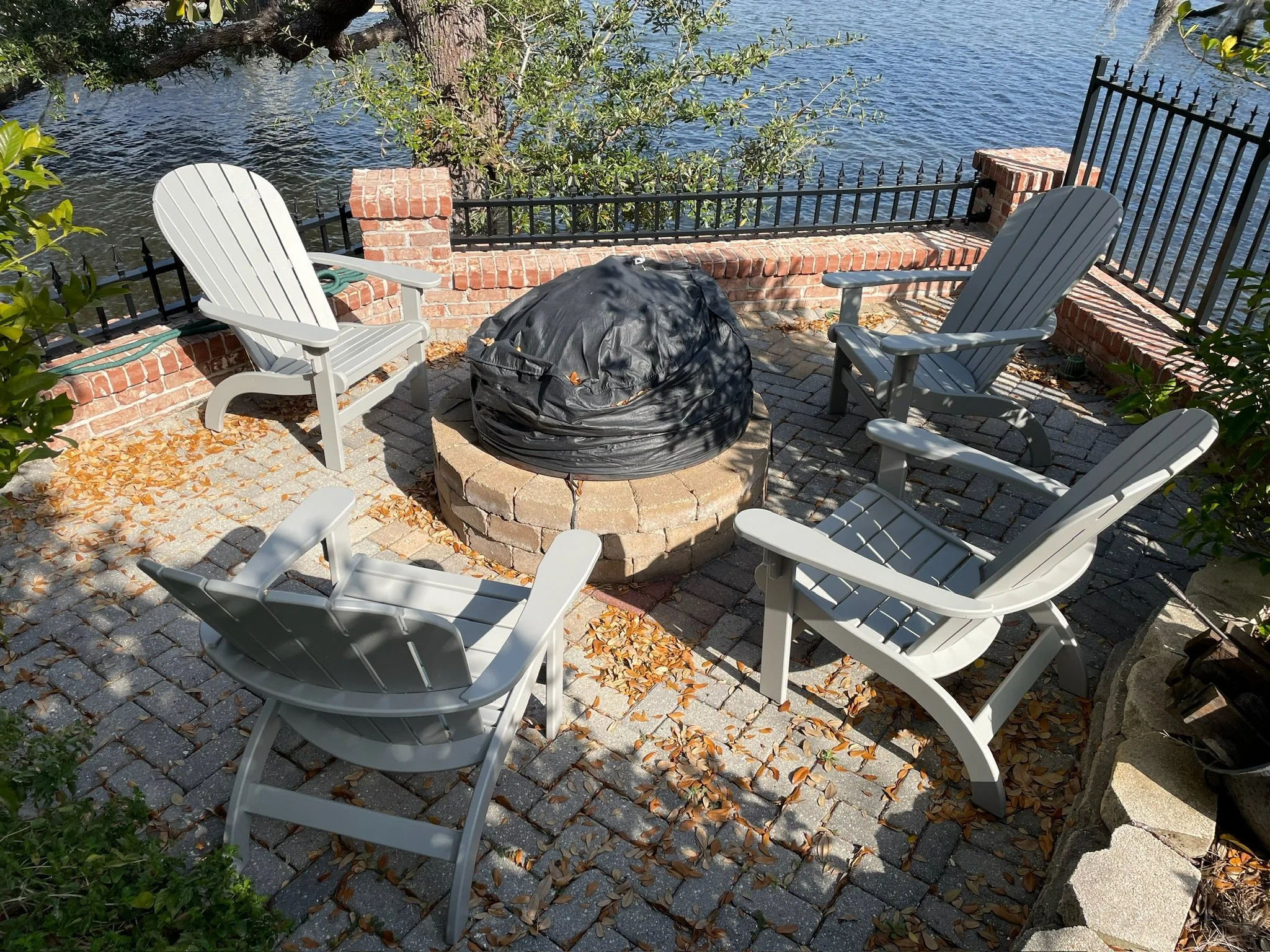 Four white Adirondack chairs arranged around a fire pit on a brick patio, with a view of a body of water and trees in the background.