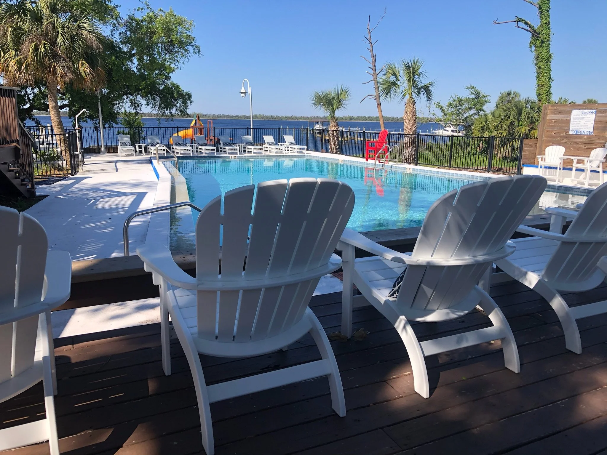 Outdoor swimming pool with lounge chairs, a play structure, and a view of the water and boats beyond a black fence. Palm trees and a clear blue sky are visible.
