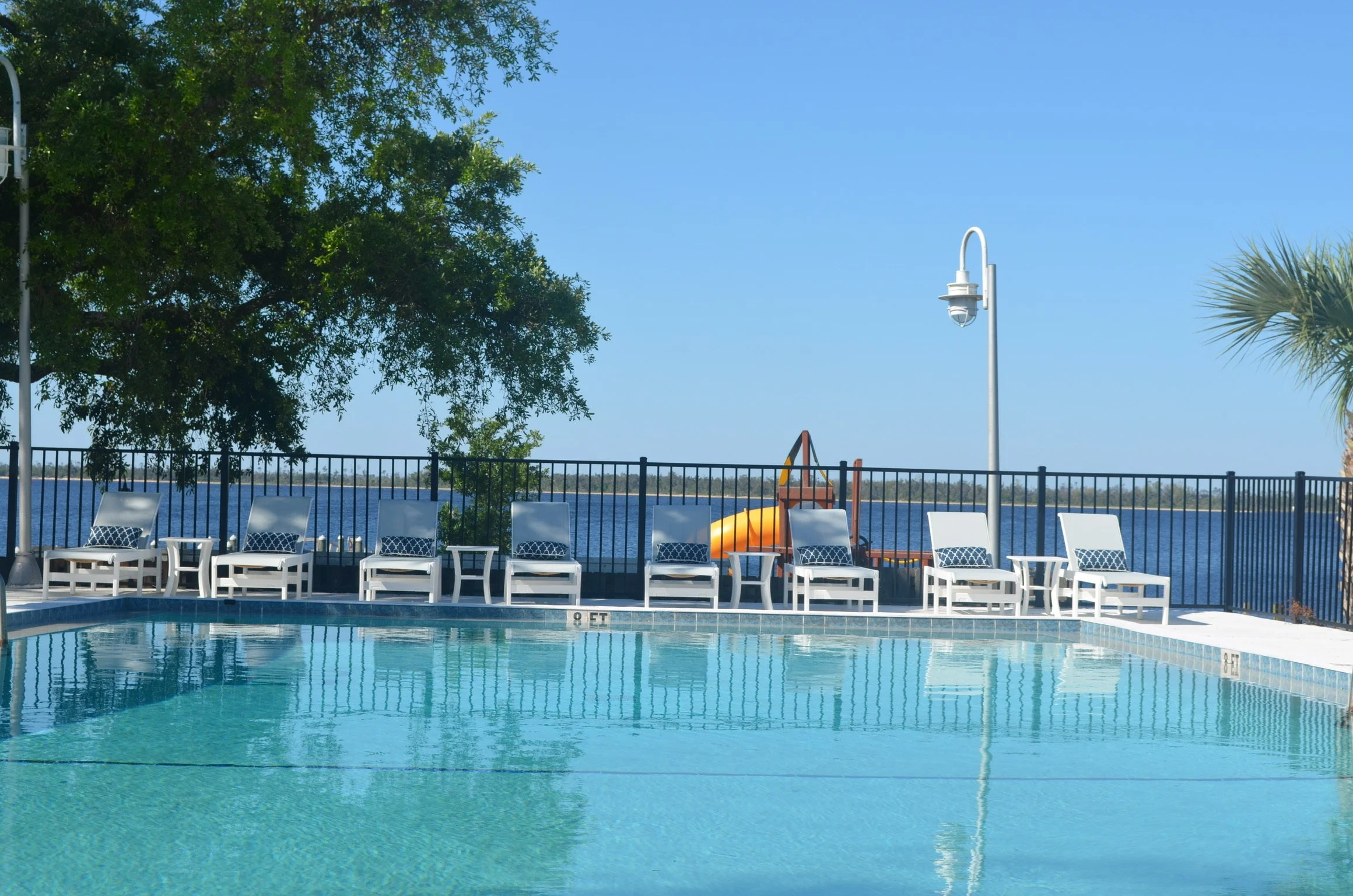 An outdoor swimming pool with lounge chairs along the edge, overlooking a water body with a tree and blue sky in the background.
