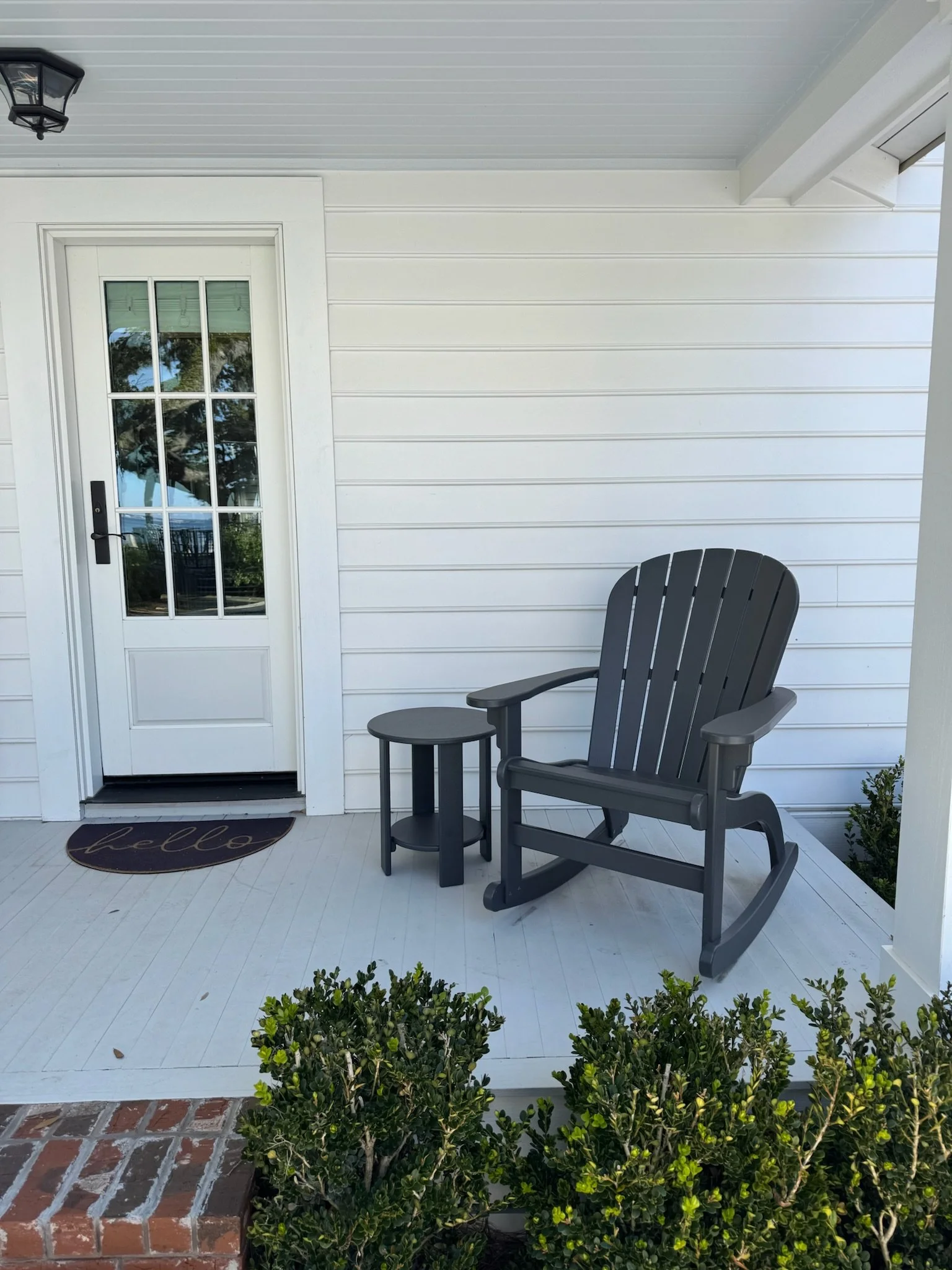 Front porch area with white siding, a glass-paneled door, a black Adirondack chair, a small black side table, a doormat that says "hello," green bushes, and brick steps.