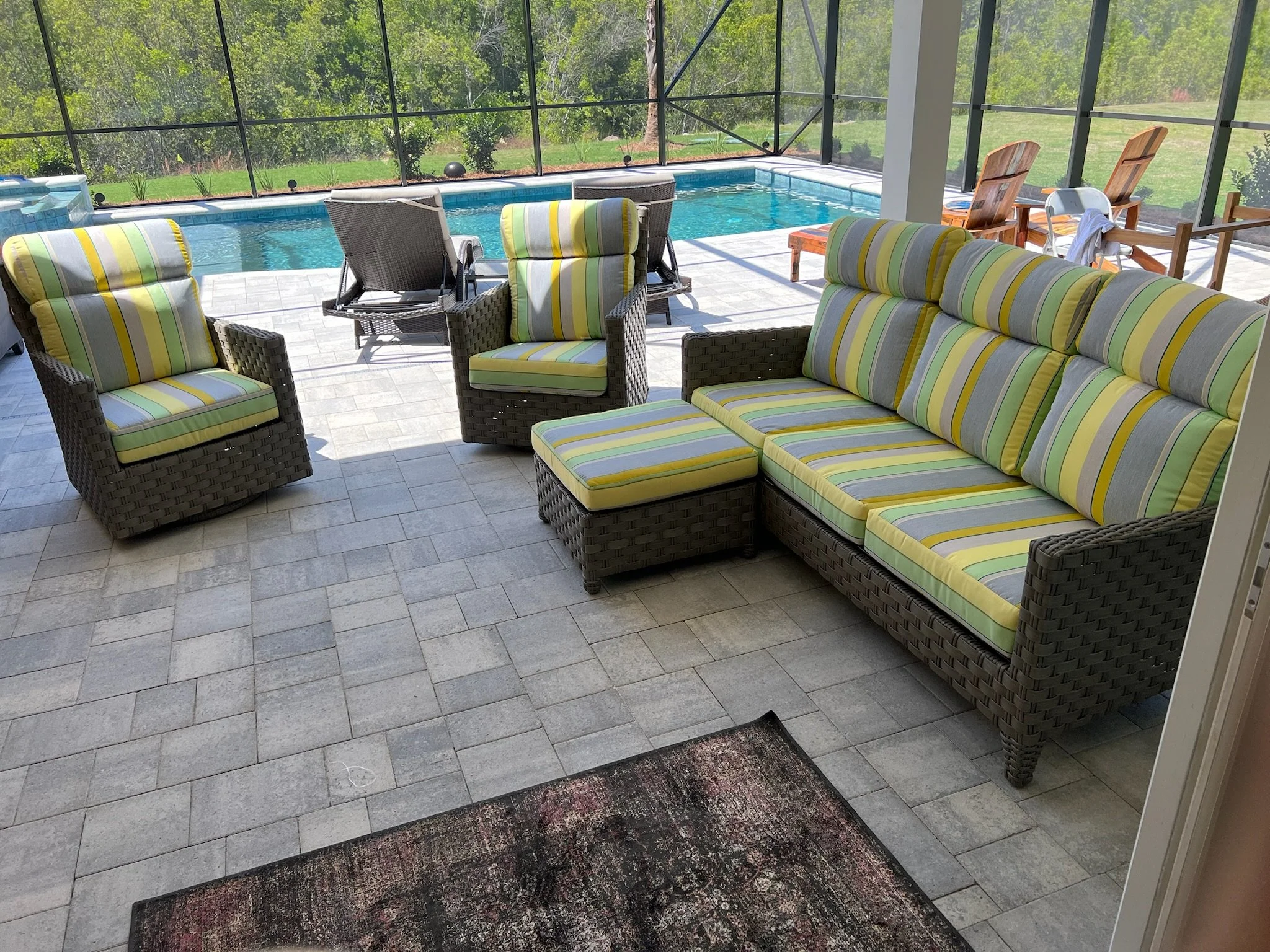 A screened-in pool area with lounge chairs, wicker chairs with striped cushions, and a table with two wooden chairs, overlooking a grassy yard and trees.