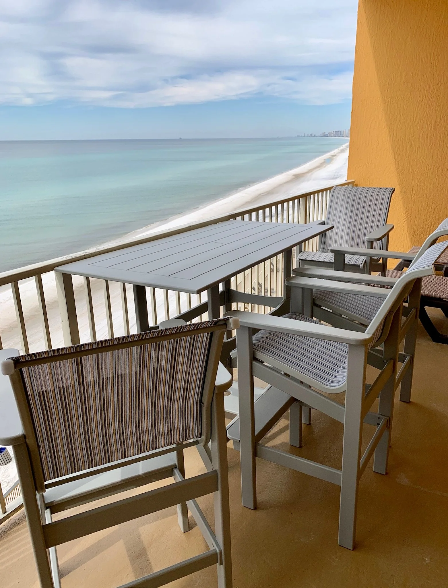 Balcony view overlooking the ocean with a table and chairs, yellow wall on the right, cloudy sky, and distant city skyline.