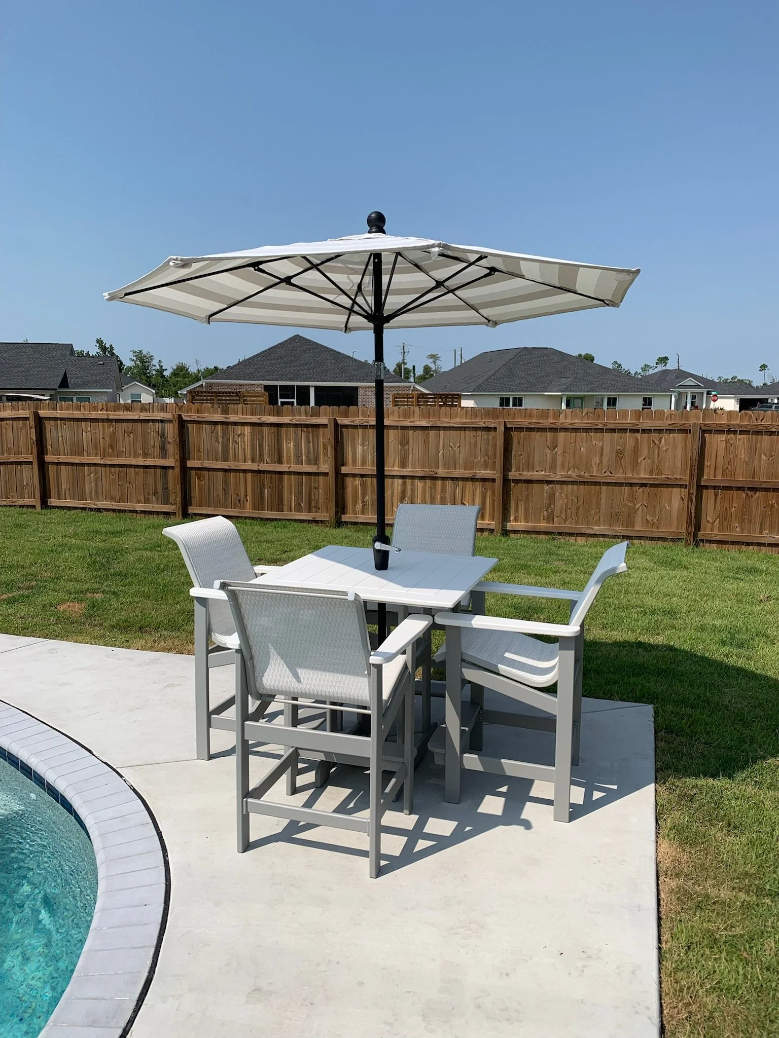 A backyard scene with a white patio table and chairs near a swimming pool, a large white striped patio umbrella, a wooden privacy fence, and neighboring homes with dark roofs under a clear blue sky.