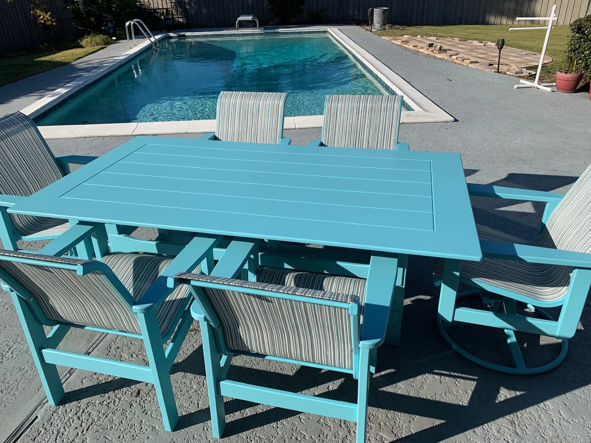 A backyard swimming pool surrounded by a concrete deck, with a light blue outdoor table and striped chairs in the foreground. There's a fenced yard with grass and some plants, and a stone patio area with a potted plant and a lawn spray hose in the ba