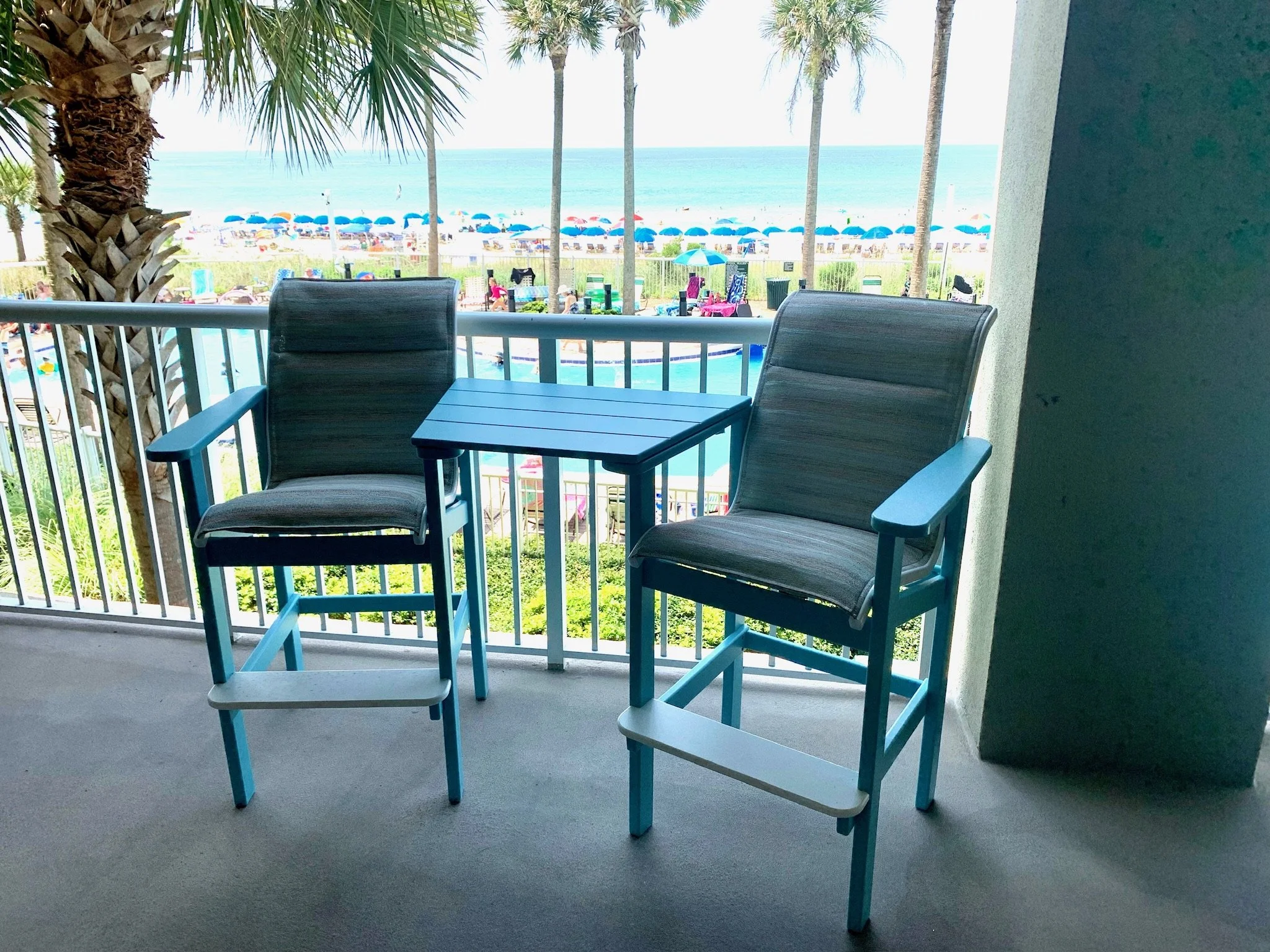 Balcony with two chairs and a small table overlooking a beach with colorful umbrellas, palm trees, and the ocean in the background.