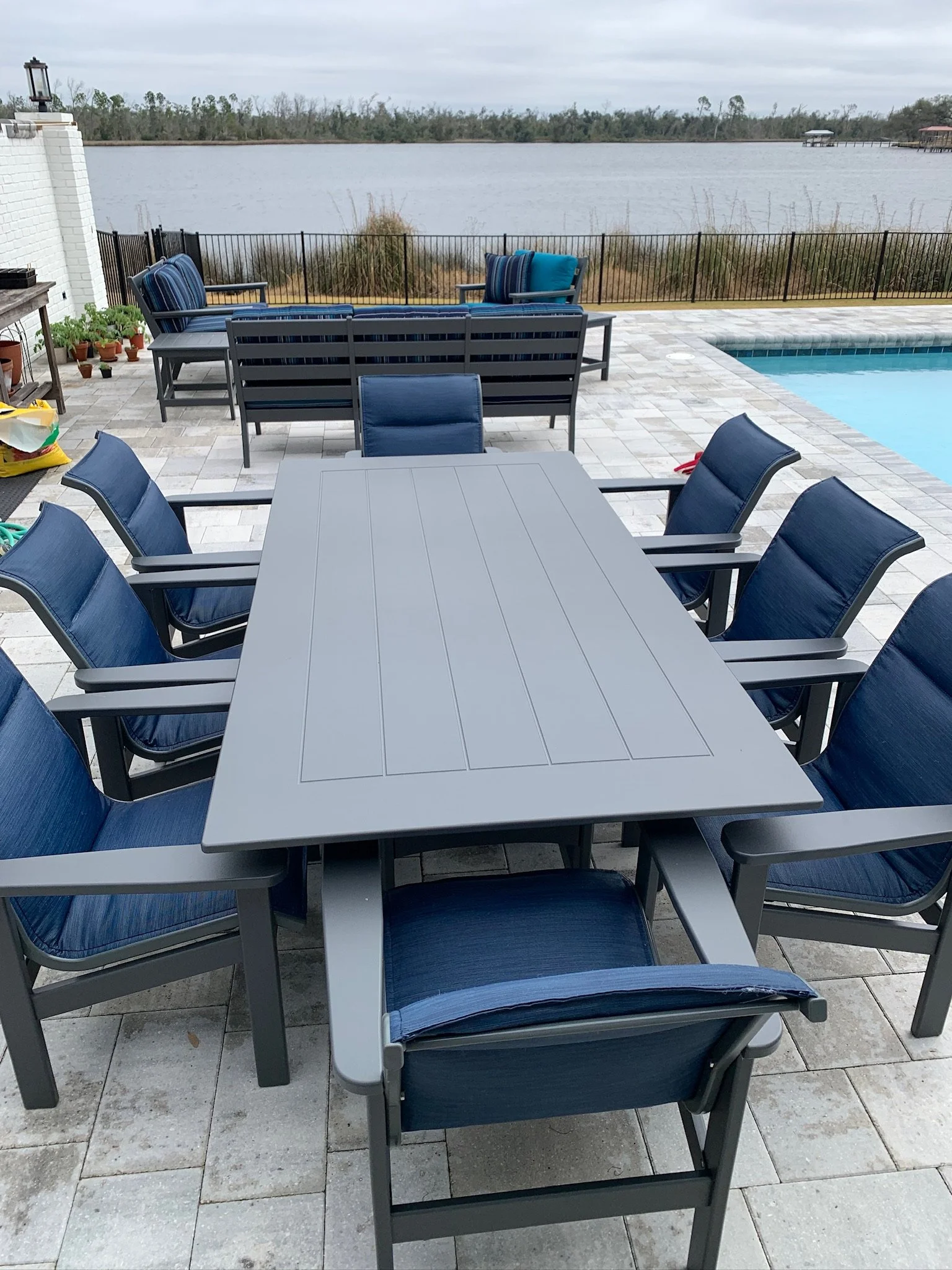 Outdoor patio with blue cushioned chairs and a dining table, overlooking a lake with a stone pool deck, plants in pots, and cloudy sky.