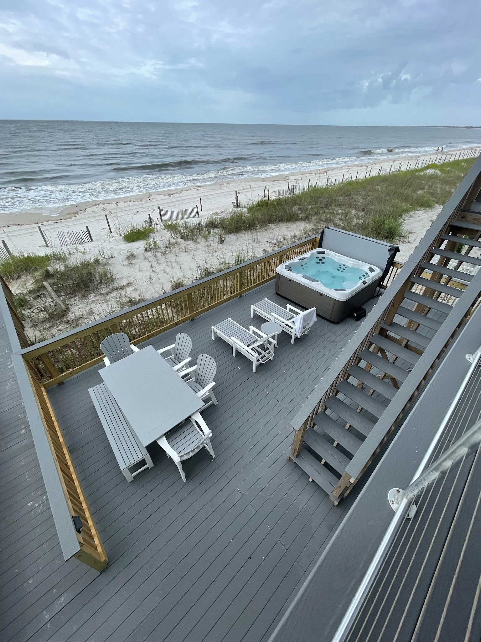 A balcony with outdoor furniture, including a dining table with chairs, a hot tub, and lounge chairs overlooking a beach with sand, grass, fencing, ocean waves, and cloudy sky.
