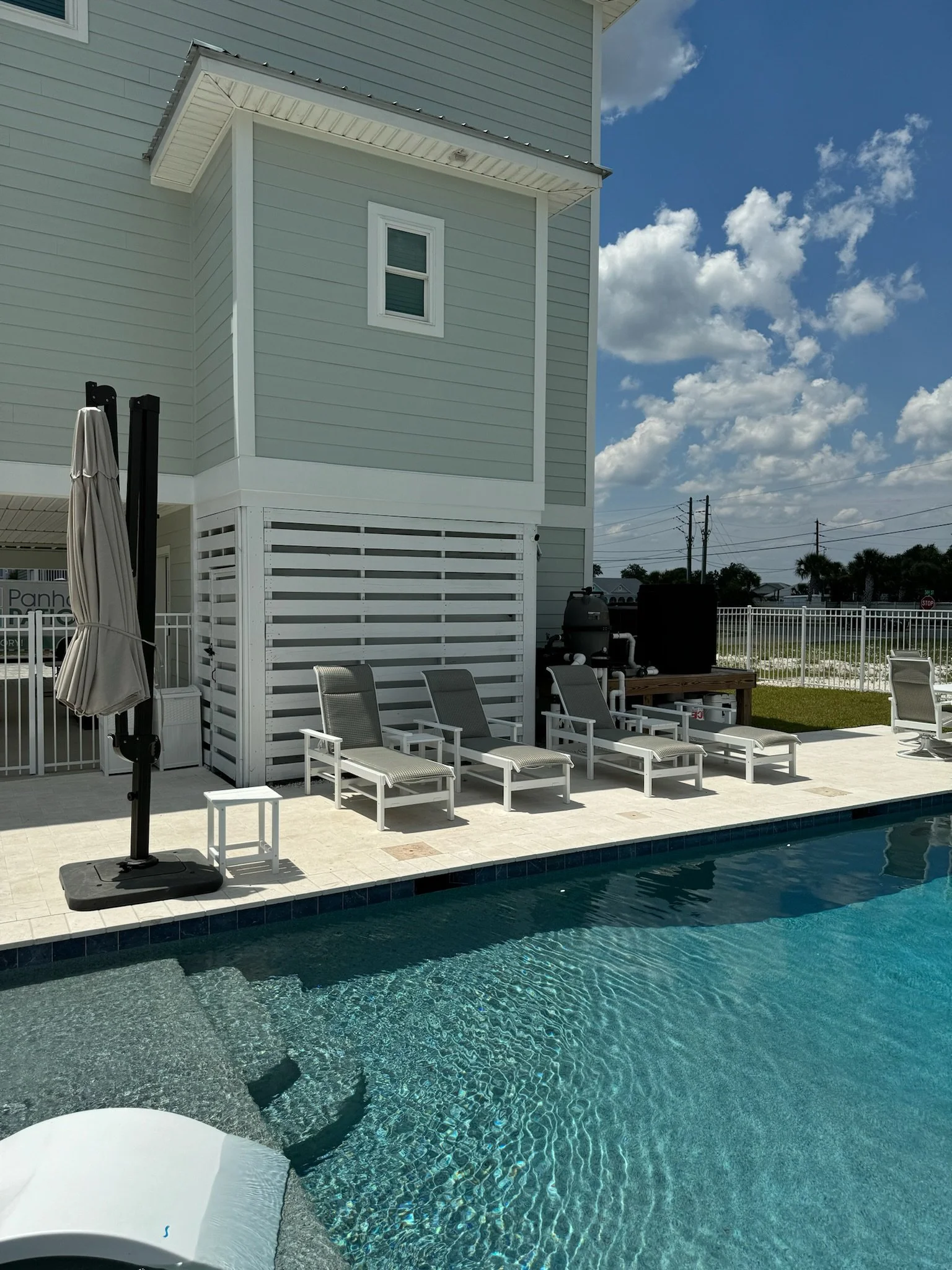 An outdoor swimming pool with clear blue water and a concrete deck area with lounge chairs. There is a white fence in the background, and an light gray house with white trim and siding. The sky is bright with some clouds.