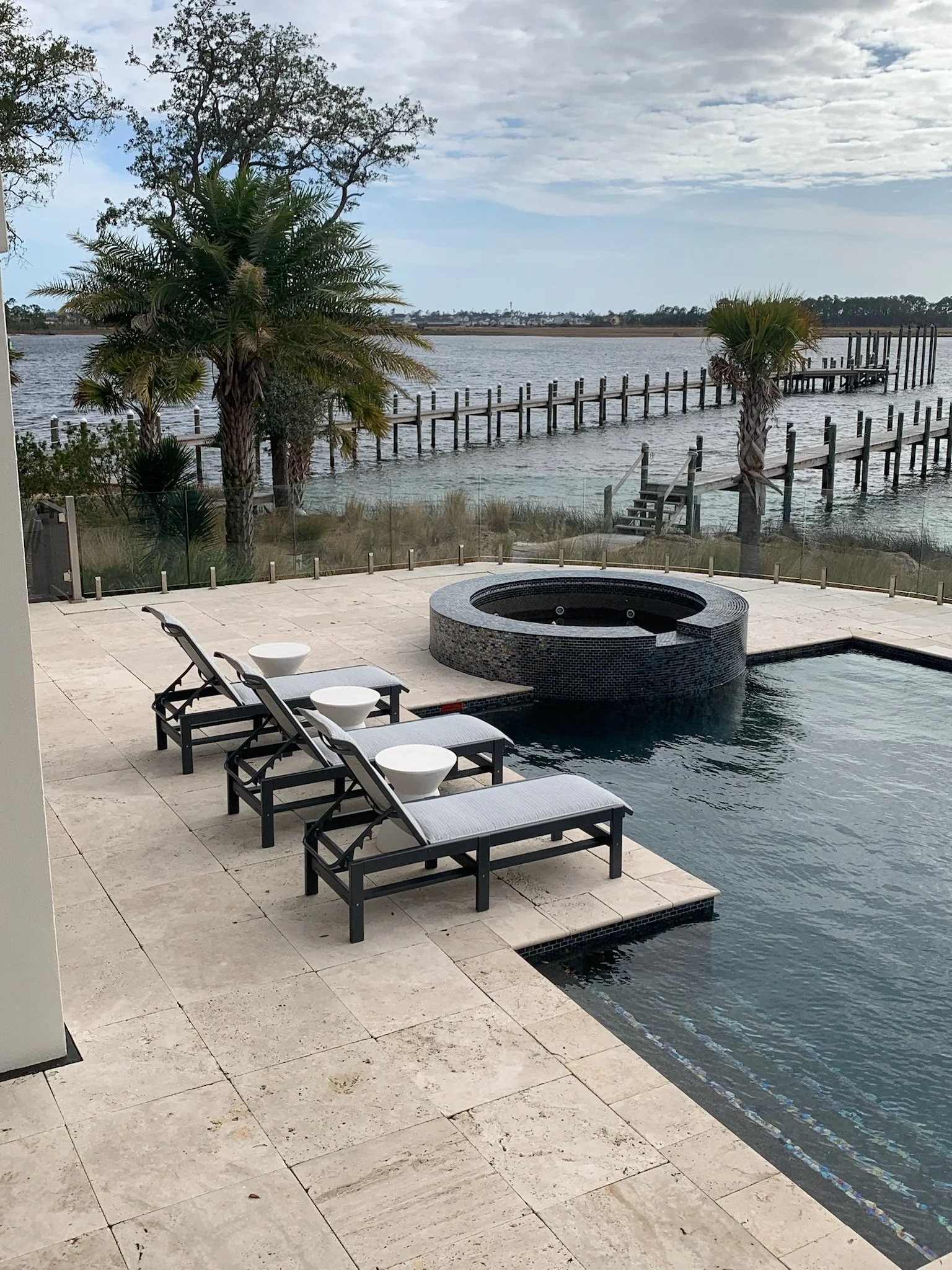 A backyard patio with lounge chairs, small round tables, a fire pit, and a view of a lake or river with a wooden dock extending into the water surrounded by palm trees and coastal vegetation. Overcast sky with patches of blue.