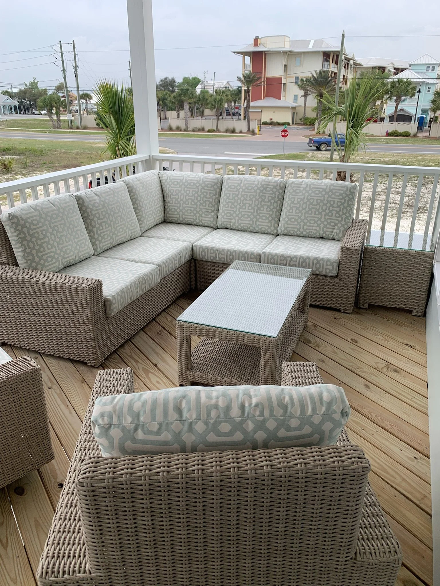 A covered porch with wicker furniture including a sofa and a coffee table, overlooking a street with palm trees and multi-story residential buildings.