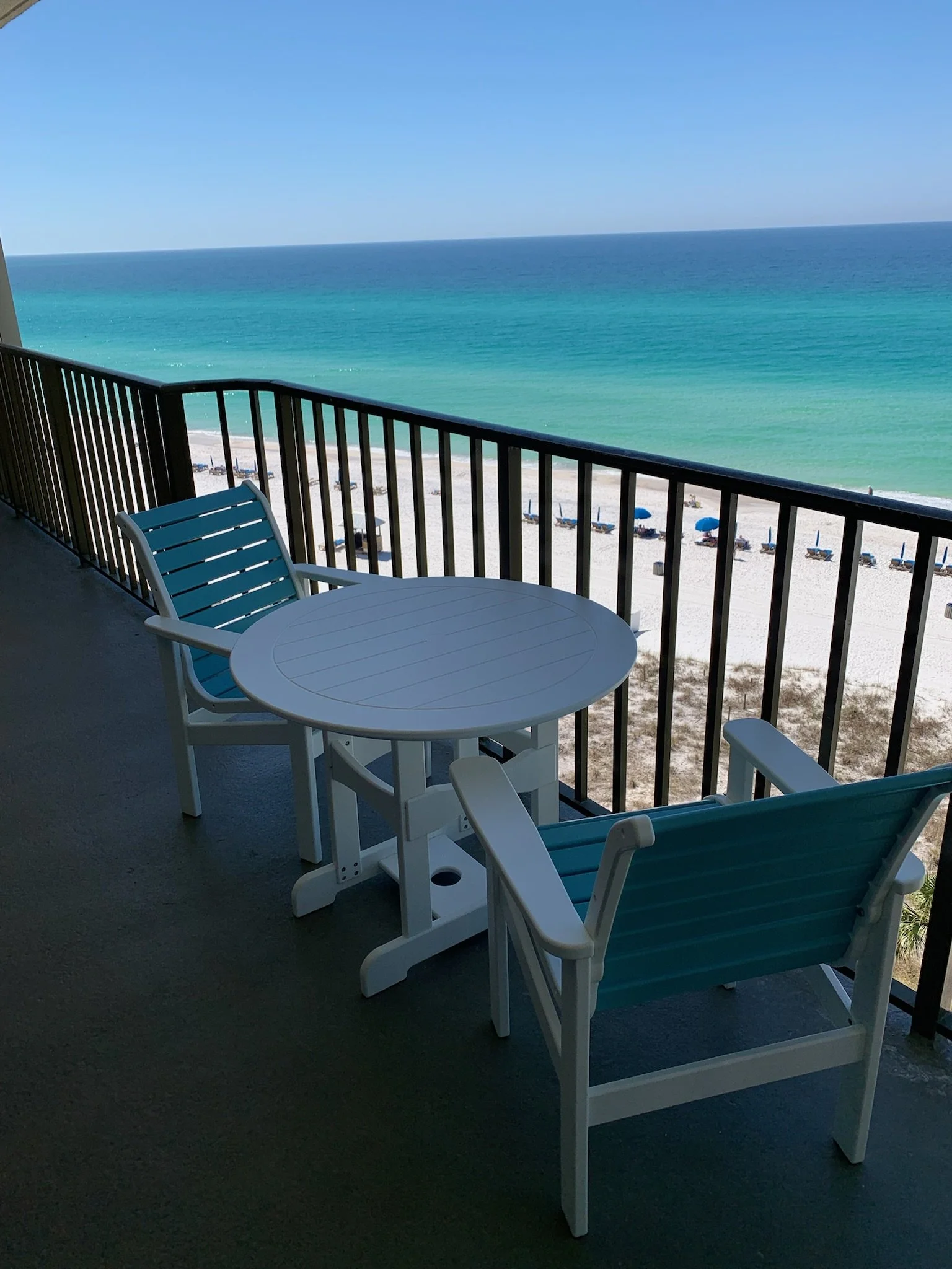 A balcony with a white round table and two blue-striped chairs overlooking a sandy beach, blue ocean, and clear sky.