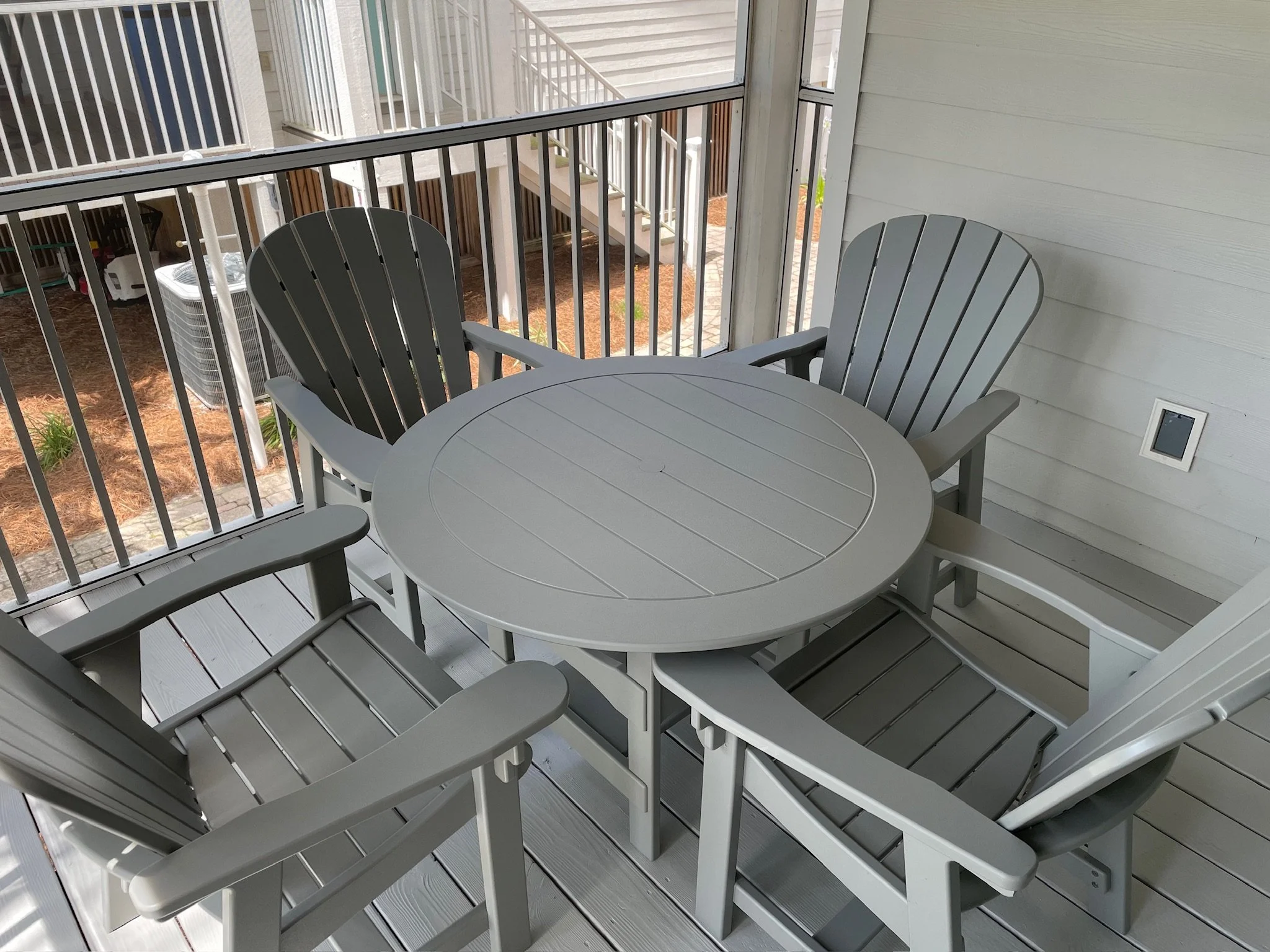 A balcony with a round table and four gray Adirondack chairs. A safety railing surrounds the balcony, and neighboring apartment stairs are visible beyond the railing.