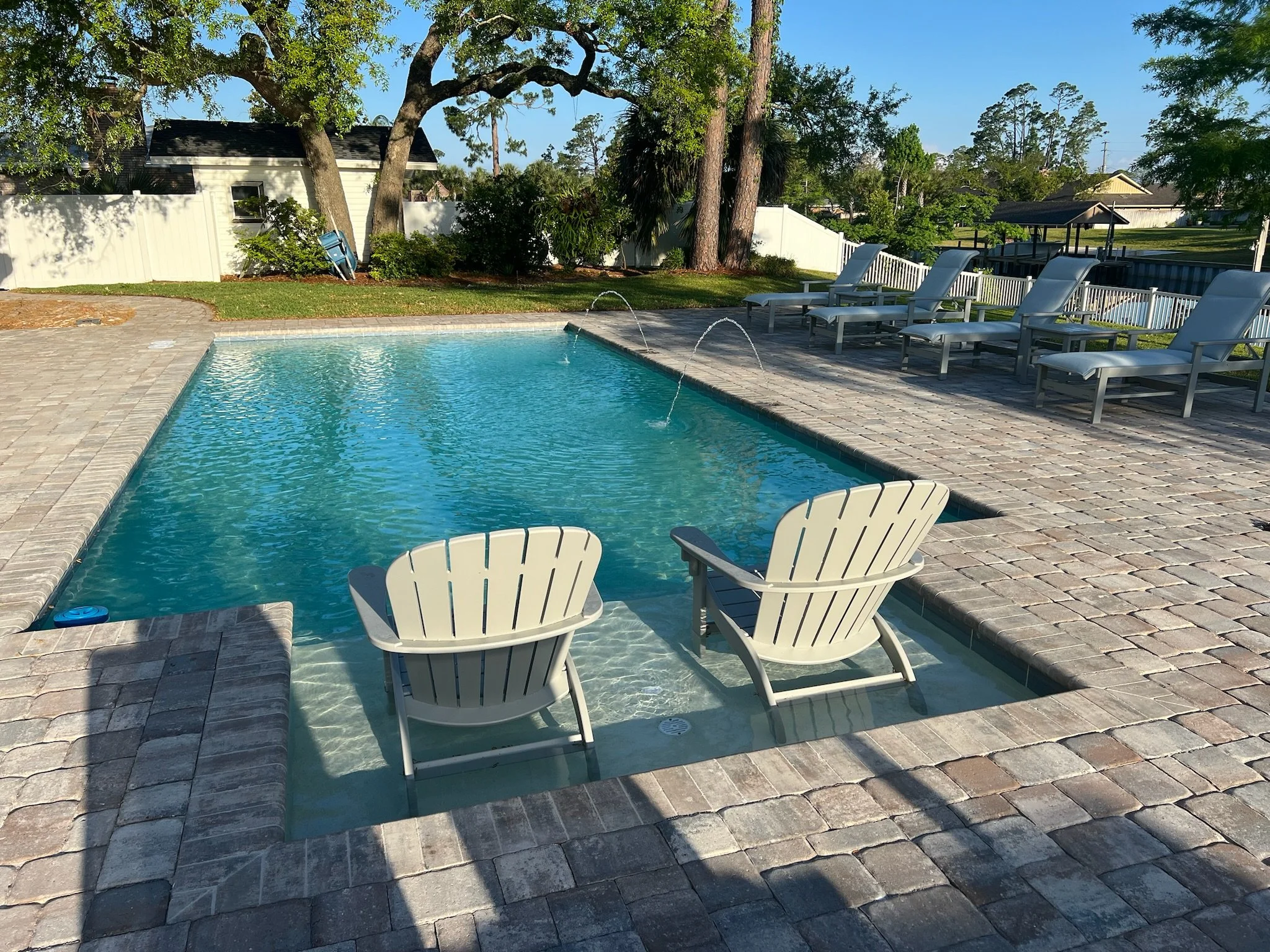 An outdoor swimming pool surrounded by a stone patio and white lounge chairs. Trees and a white vinyl fence enclose the backyard.