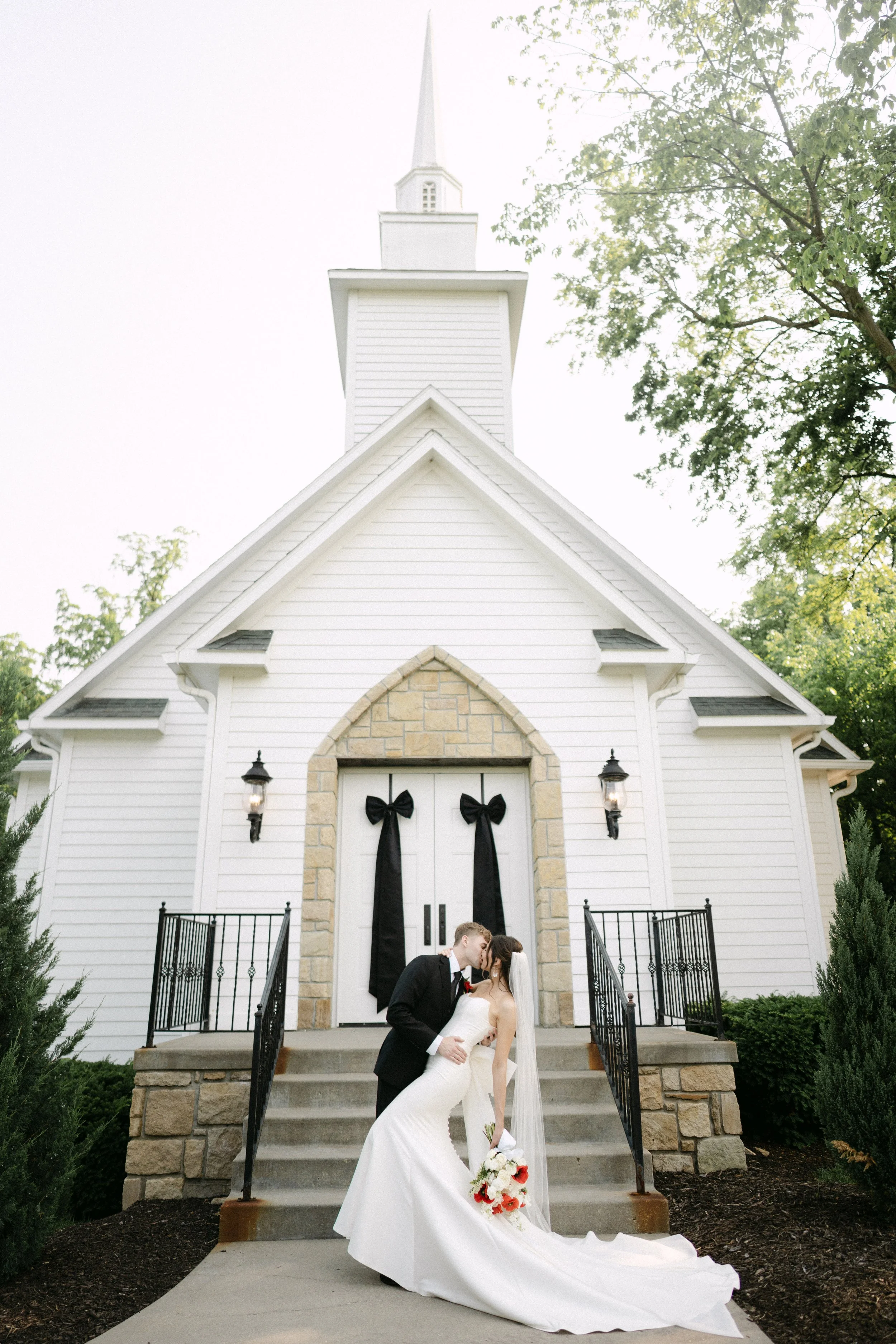A newlywed couple sharing a kiss in front of a white church with a tall steeple during daytime. The bride is in a strapless wedding gown holding a bouquet of white and red flowers, and the groom is in a black tuxedo. The church has black bow decorations on its doors and black lantern-style lights on either side.