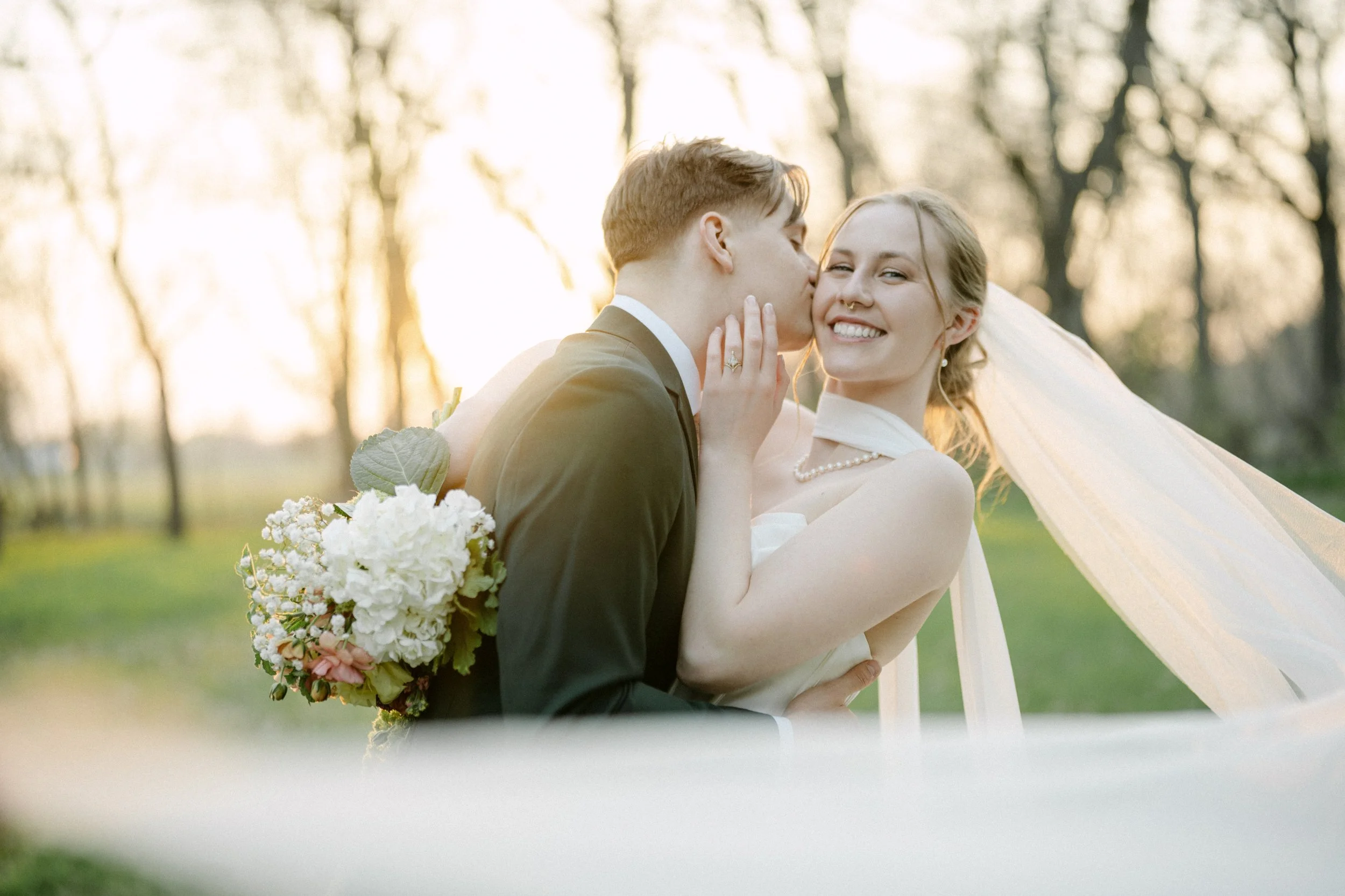 A newlywed couple in wedding attire sharing a tender moment outdoors during sunset, with the groom kissing the bride on the cheek and the bride smiling happily, holding a bouquet of white and pink flowers.