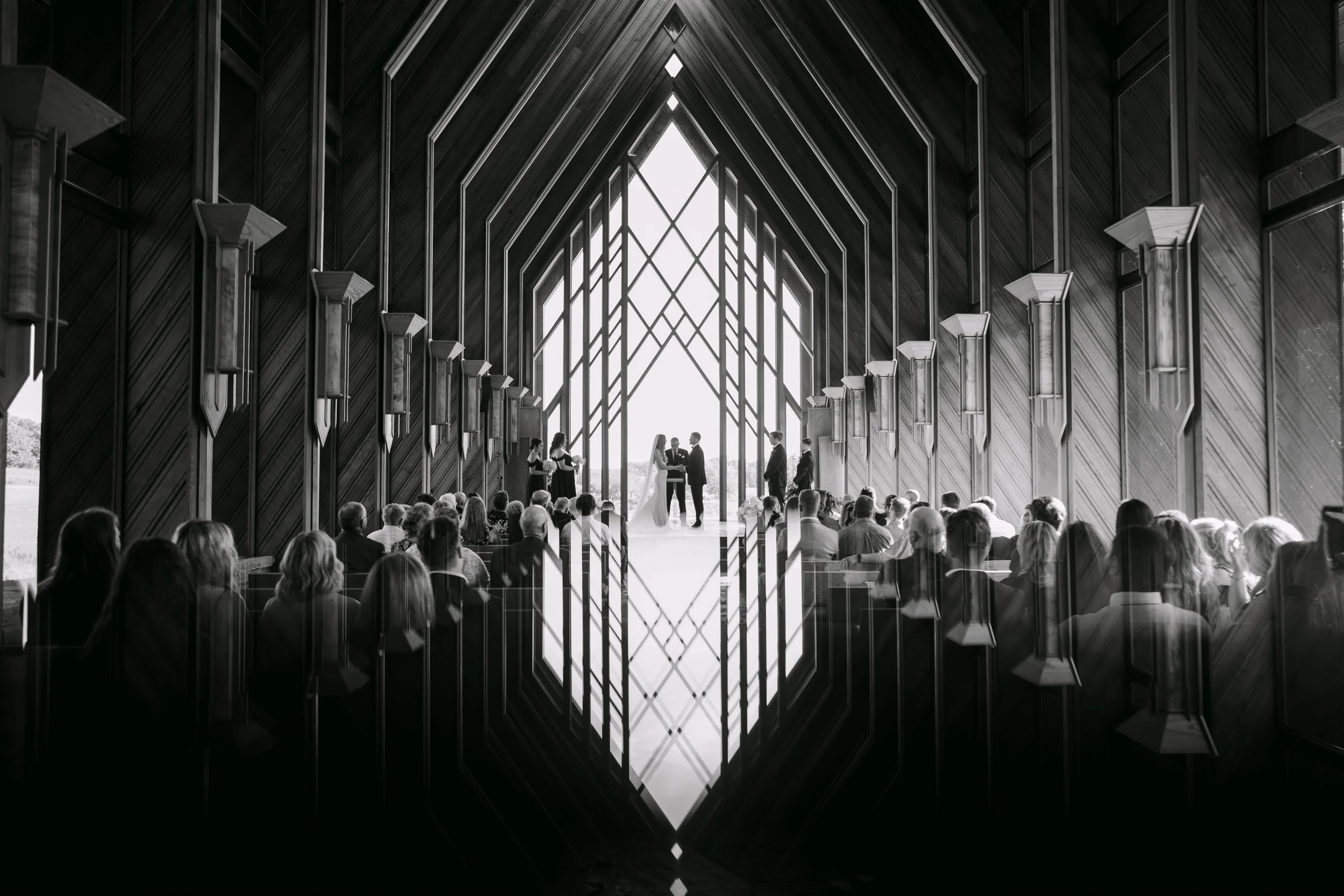 A wedding ceremony taking place inside a modern wooden chapel with a high, pointed ceiling and large windows. The bride and groom are standing at the altar, with the officiant and two bridesmaids nearby. Guests are seated facing the ceremony.