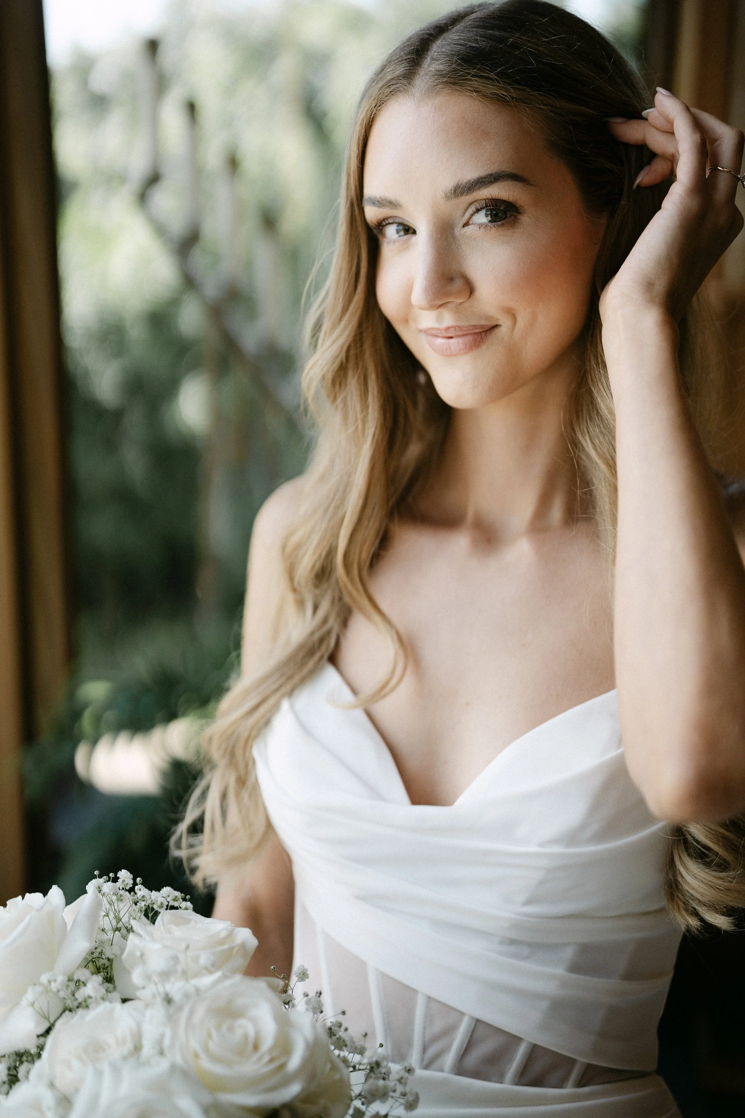 A woman with long, wavy blonde hair and blue eyes, smiling softly, is wearing a white strapless dress and adjusting her hair with her right hand. She is standing in front of a window with greenery outside, and a bouquet of white flowers is in the foreground.