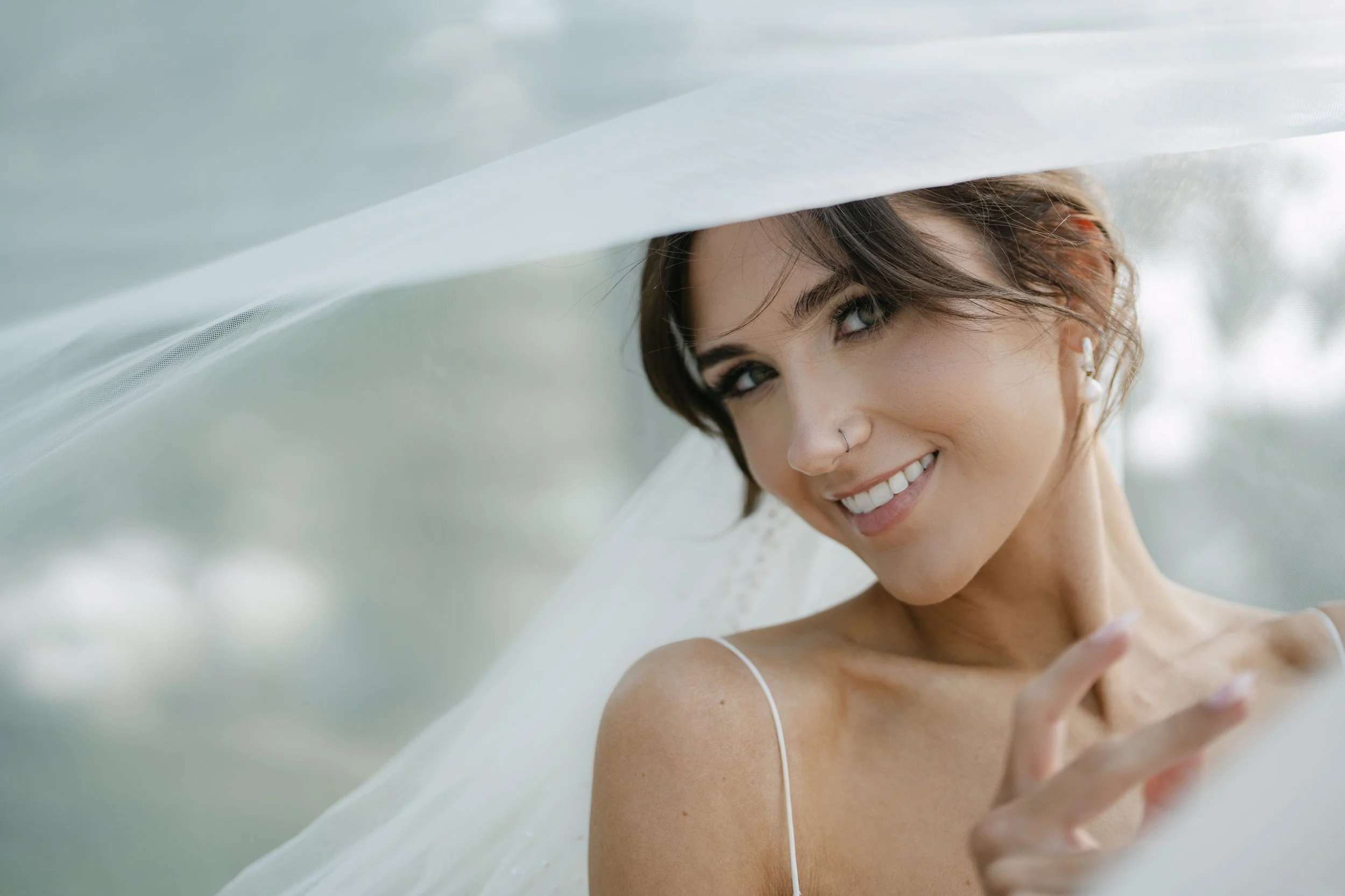 Close-up of a smiling woman with short brown hair, wearing a white spaghetti strap top, and looking up with a white sheer fabric partially covering her face.