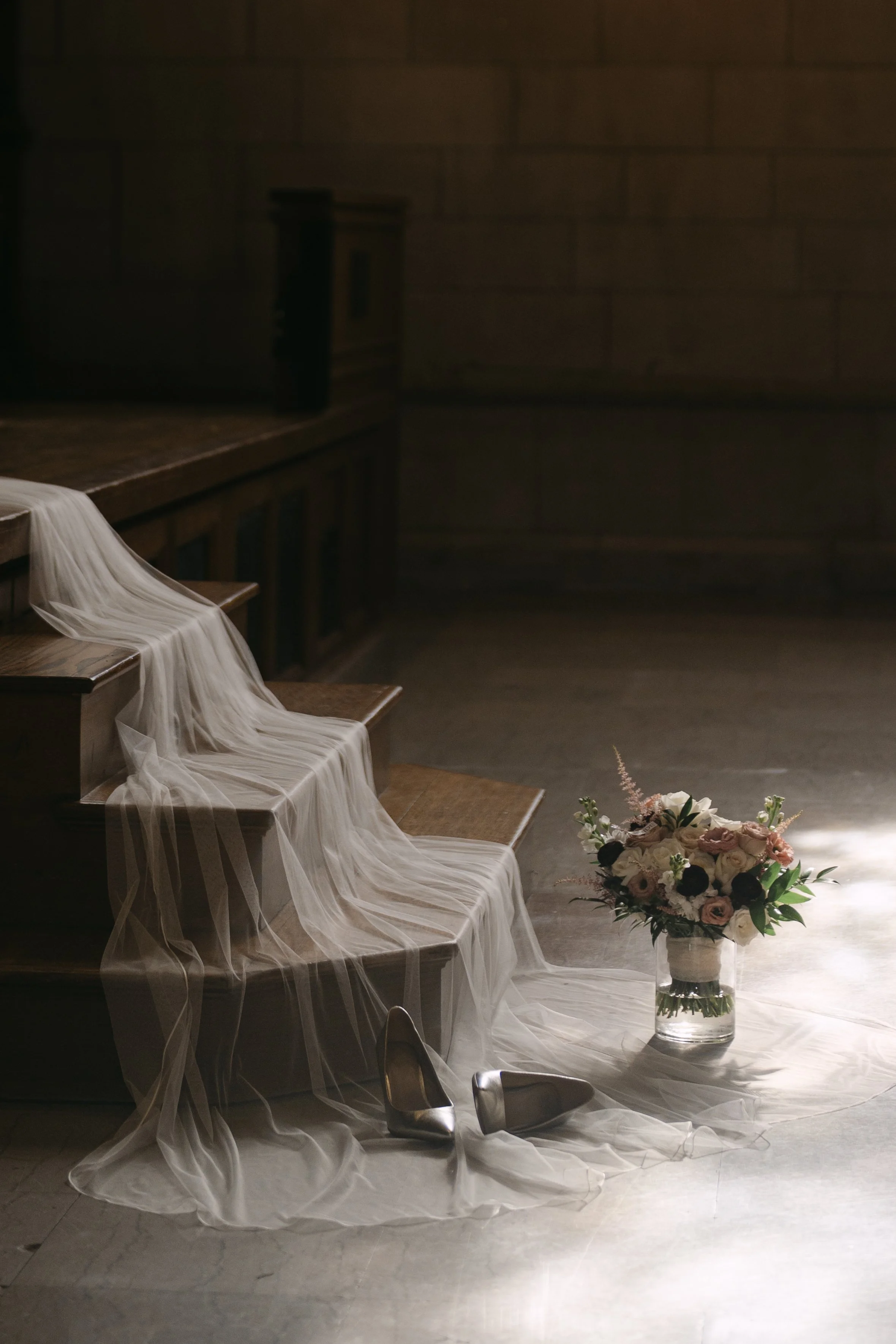 A bouquet of flowers in a glass vase placed on the floor, with sheer fabric draped over wooden stairs, a wooden cabinet, and a brick wall in the background, suggesting a romantic or wedding decor setting.