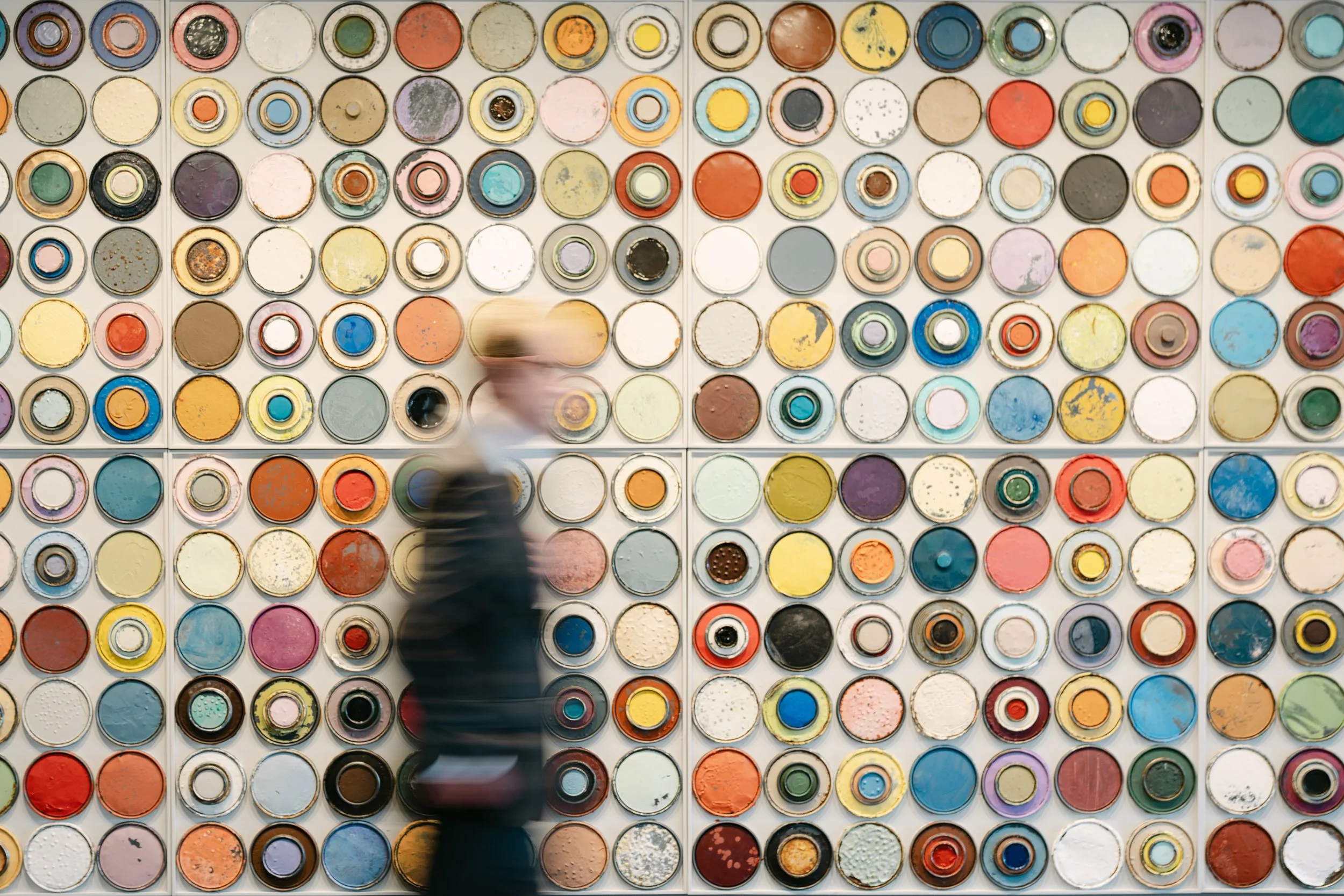 Colorful cans of paint arranged neatly on a wall, with a person walking past, seen as a blurry figure.