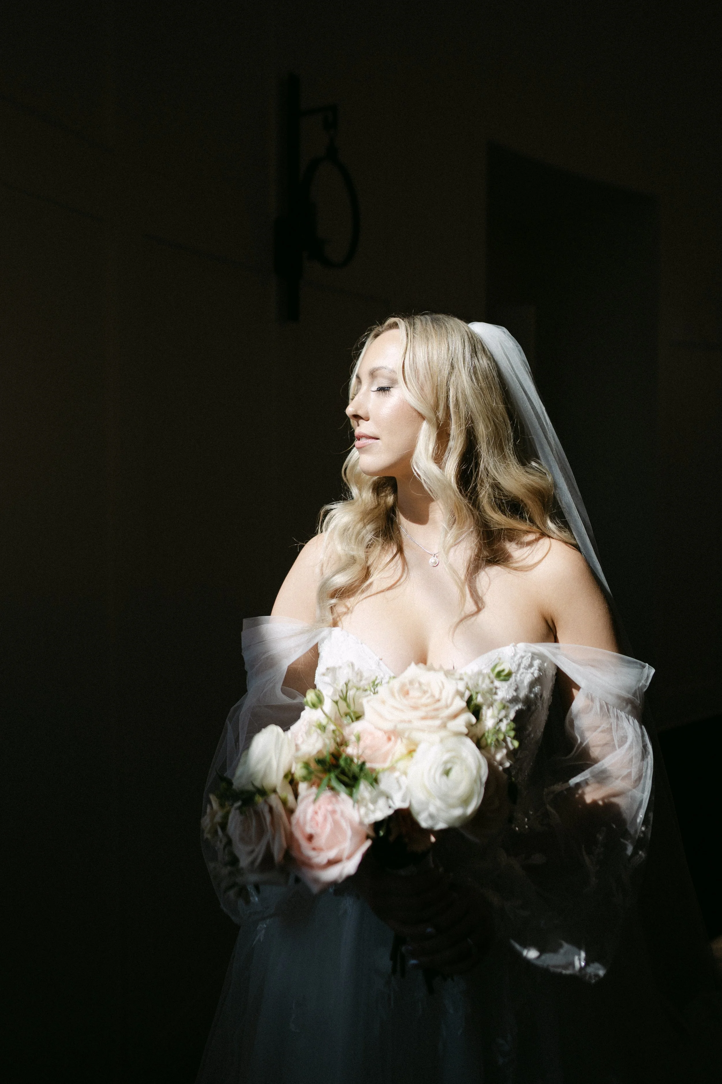 A bride with long blonde hair, closed eyes, wearing a wedding dress and a veil, holding a bouquet of white, blush pink, and pale peach roses, against a dark background.