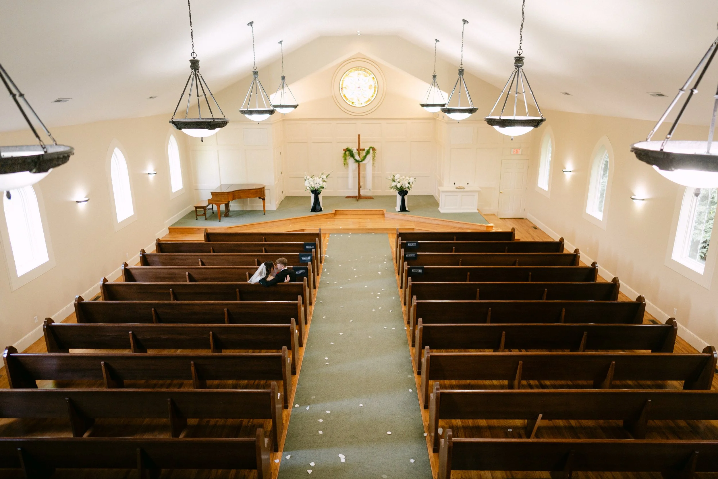 Interior of a church with wooden pews, a green aisle runner, and a wedding couple sitting together. The altar has floral arrangements, a cross, and a stained glass window above.