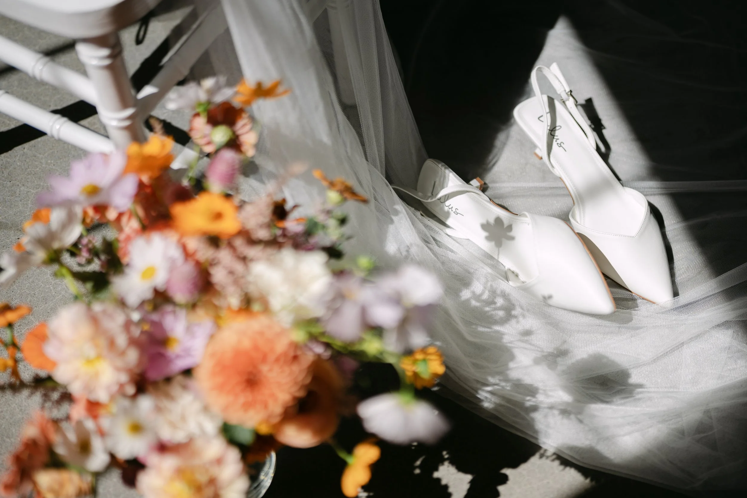 Close-up of a blurry bouquet of colorful flowers with a pair of white high-heeled shoes nearby, set on a surface with a sheer fabric and a dark backdrop.