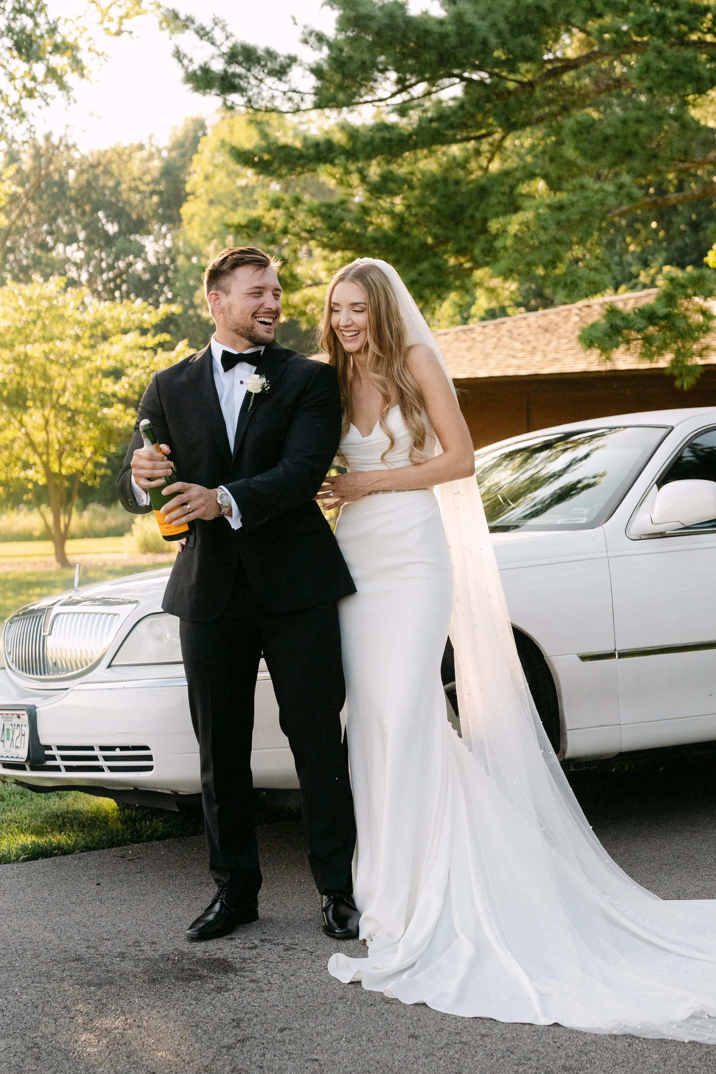 A newlywed couple standing beside a white luxury car outside, smiling and sharing a joyful moment. The man is wearing a black tuxedo with a bow tie, holding a bottle of champagne. The woman is in a white wedding gown with a long train and veil, laugh