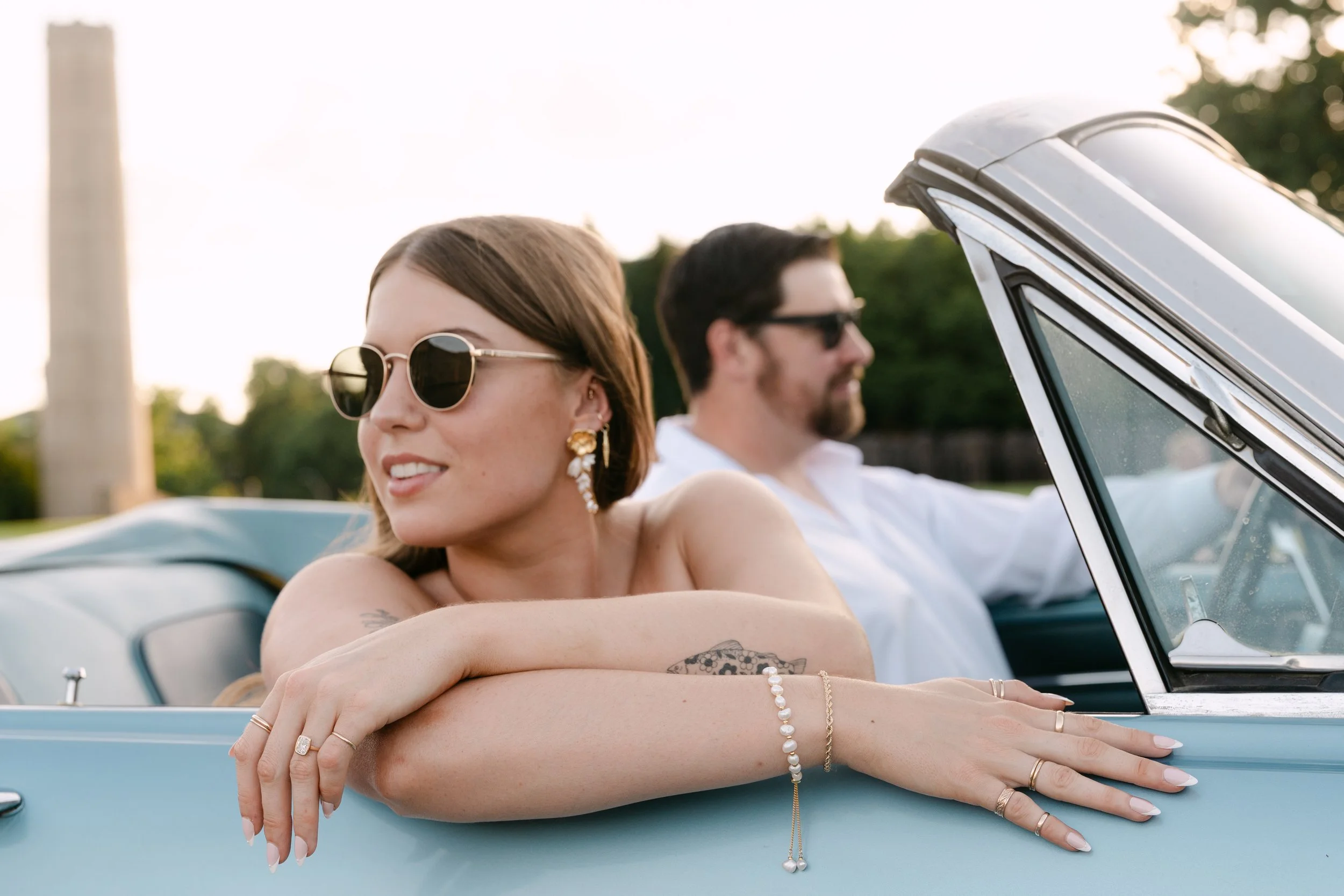 A woman with sunglasses and jewelry leaning on the side of a light blue convertible car, with a man in sunglasses in the background, both appear relaxed outdoors.