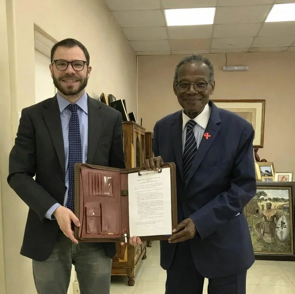 Two men in suits standing inside a room with beige walls and holding a framed document together, smiling at the camera.