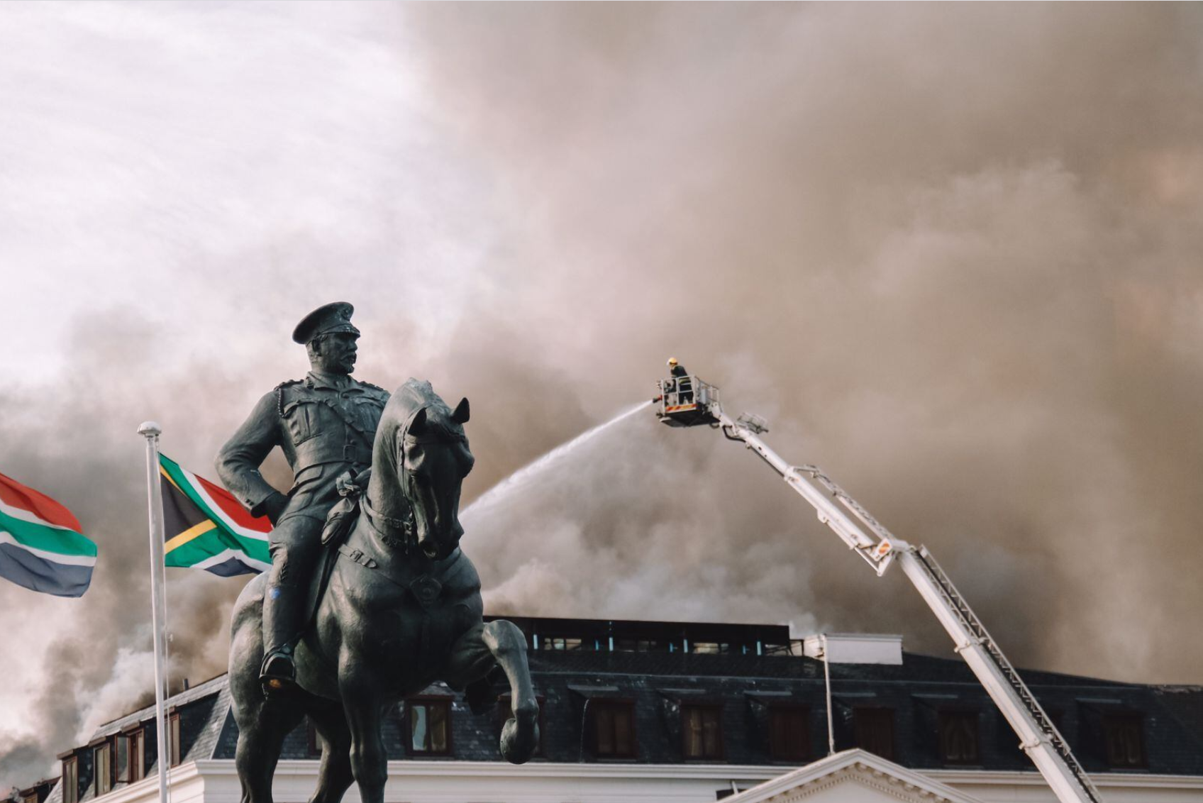 A firefighter on an extended ladder spraying water onto thick smoke in the sky above a statue of a person on horseback, with South African flags nearby.