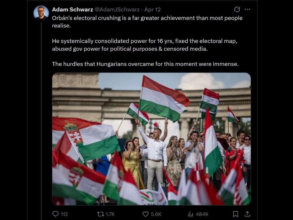 People gathered outdoors during a rally with multiple Hungarian flags waving, in front of an architectural structure, under a partly cloudy sky.