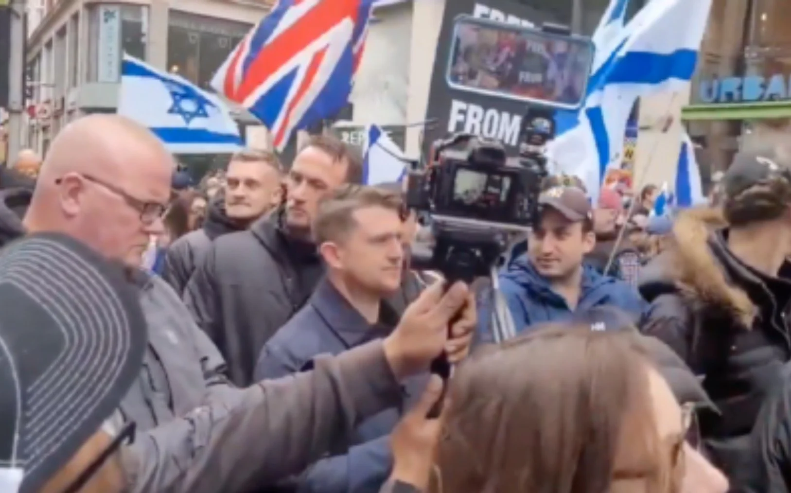 Crowd of people at a protest or rally with Israeli flags and a Union Jack flag in an urban area.
