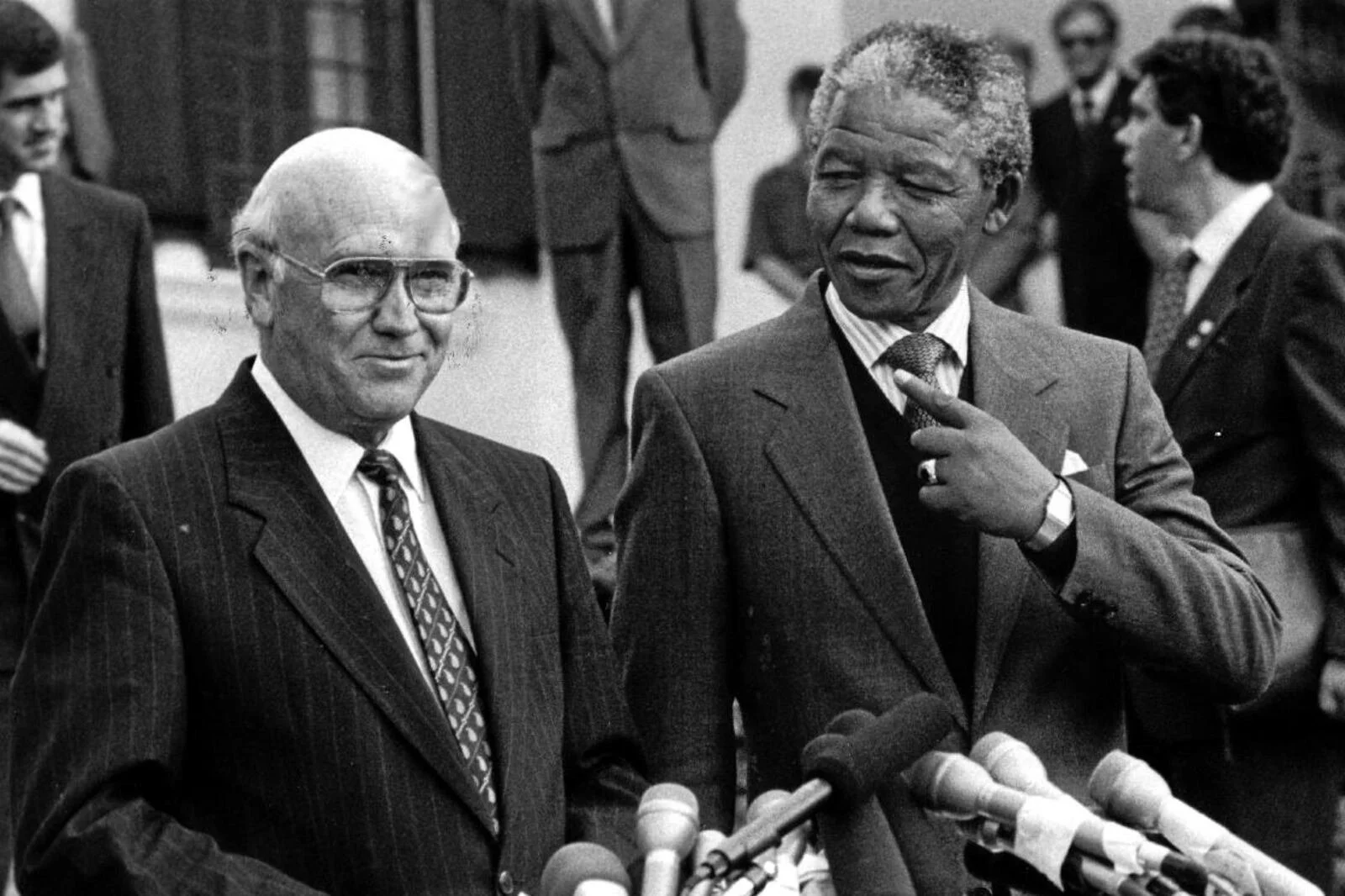 Two men in suits speaking to the media, surrounded by microphones, with other men in suits in the background.