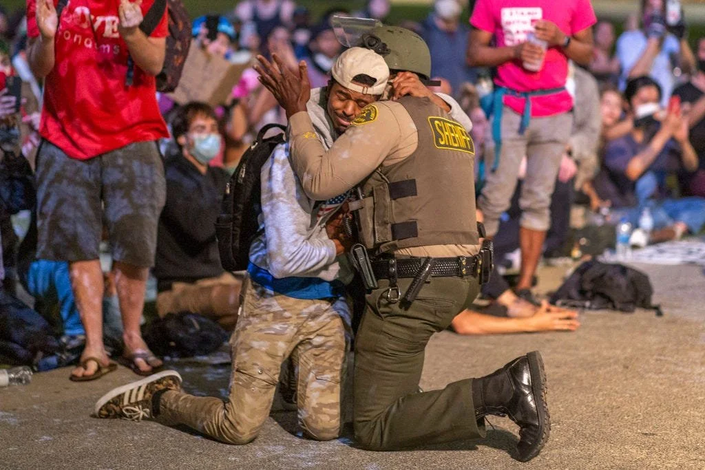 A person in camouflage clothing kneels on the ground, hugging a sheriff's deputy in uniform, amidst a crowd of onlookers. The crowd appears emotional, with some people wearing masks.