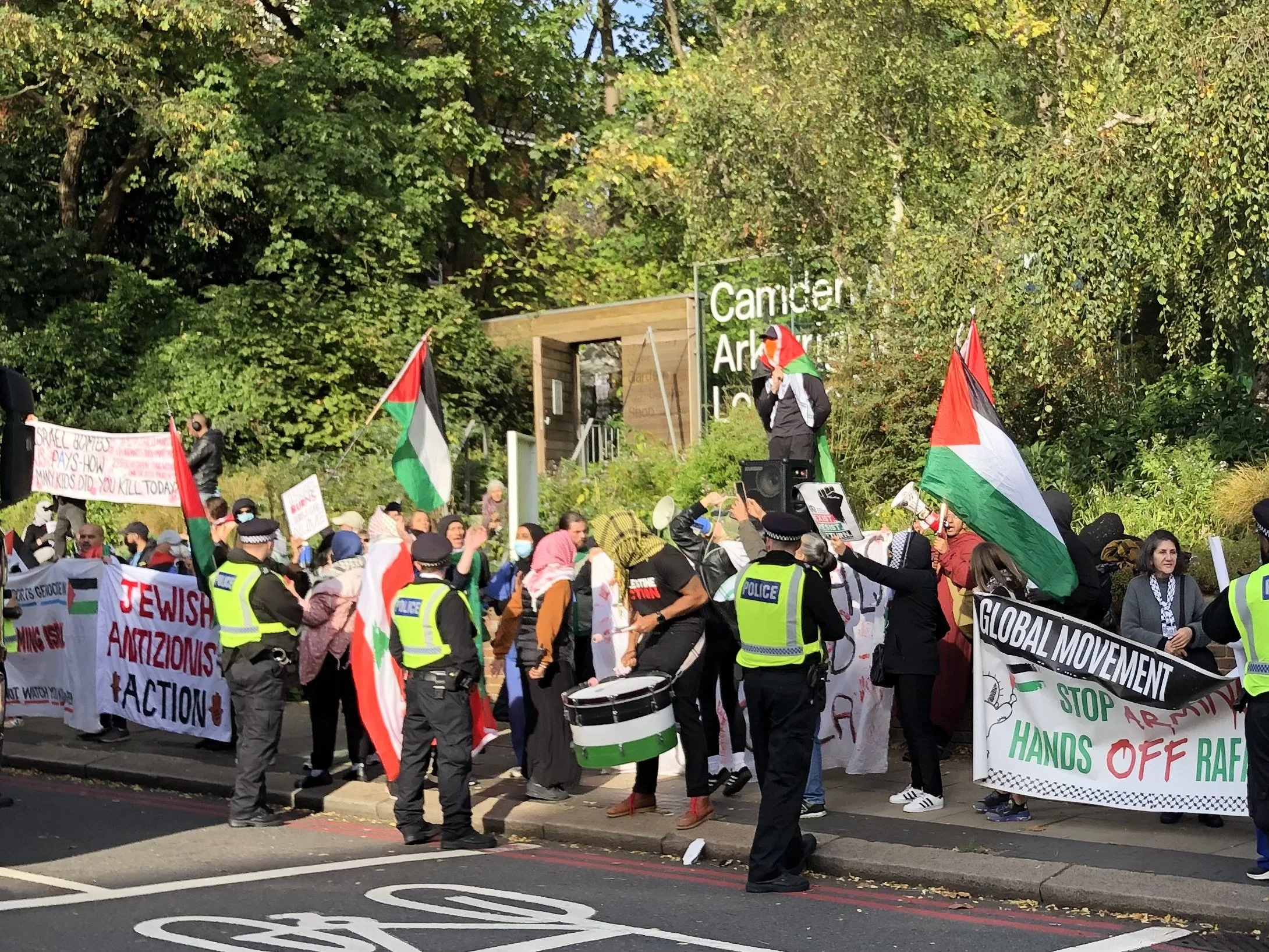A protest or demonstration with a crowd of people holding signs and Palestinian flags, with police officers present in the foreground. Trees and a sign that reads 'Canning' are visible in the background.