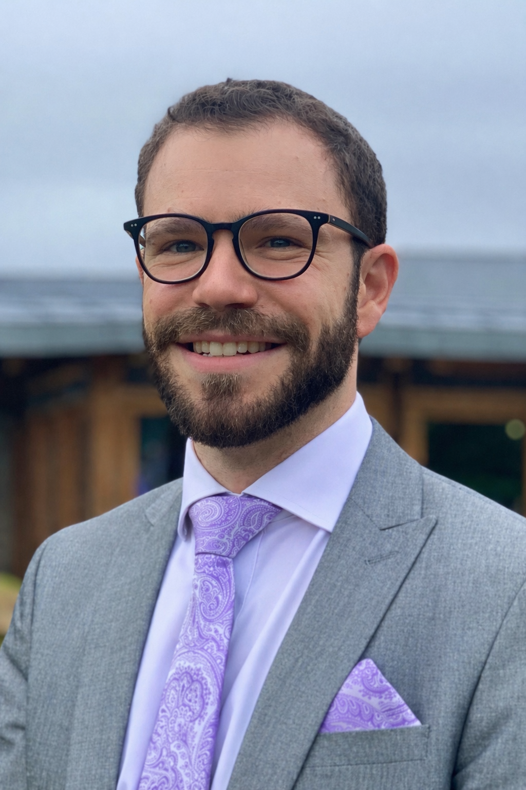 A smiling man with glasses and a beard wearing a gray suit, white shirt, lavender tie, and lavender pocket square, standing outdoors with a cloudy sky and wooden structure in the background.