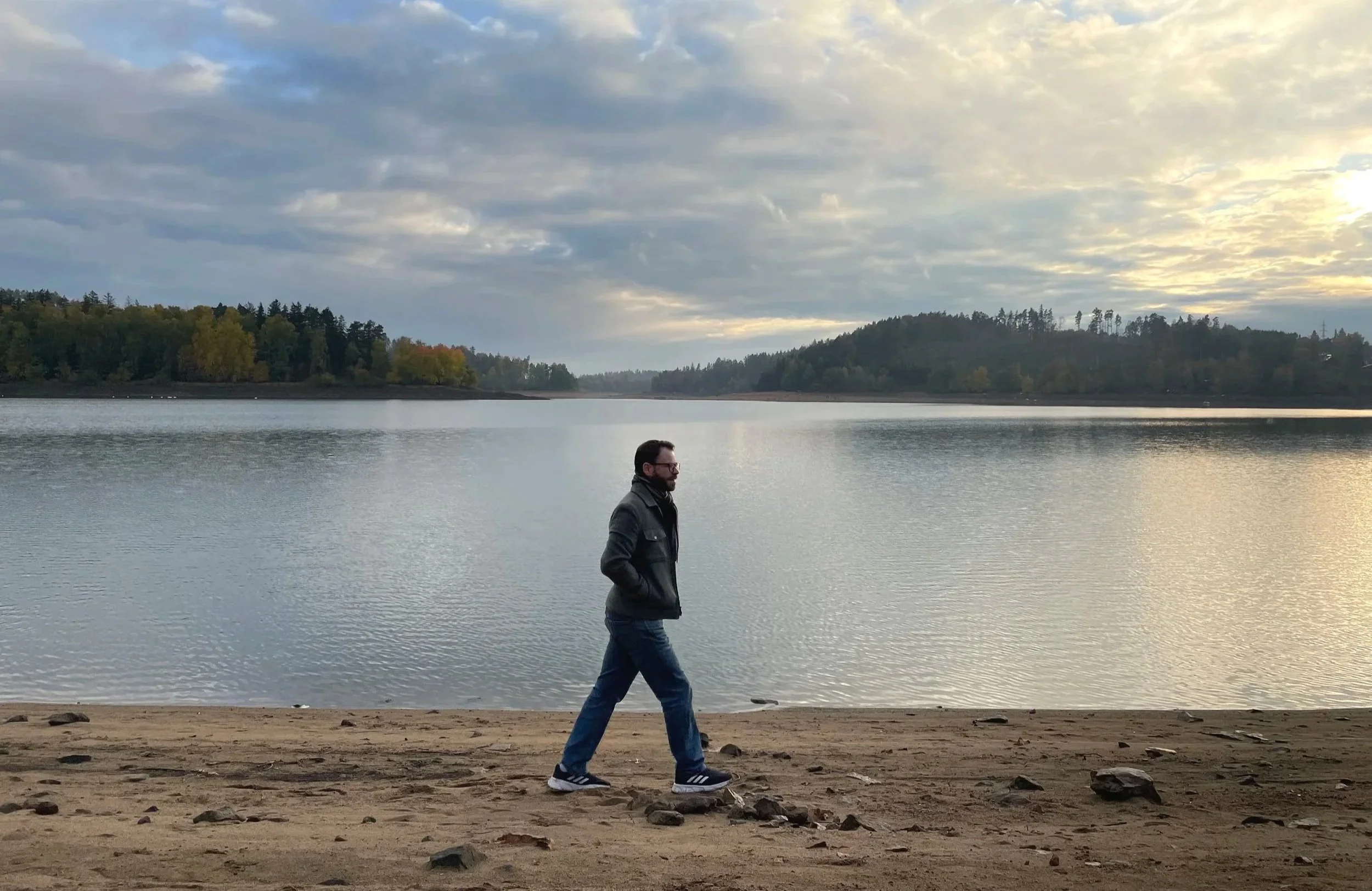 A man walking along a sandy shoreline near a calm body of water with trees on the distant hills and a cloudy sky overhead during sunset or sunrise.