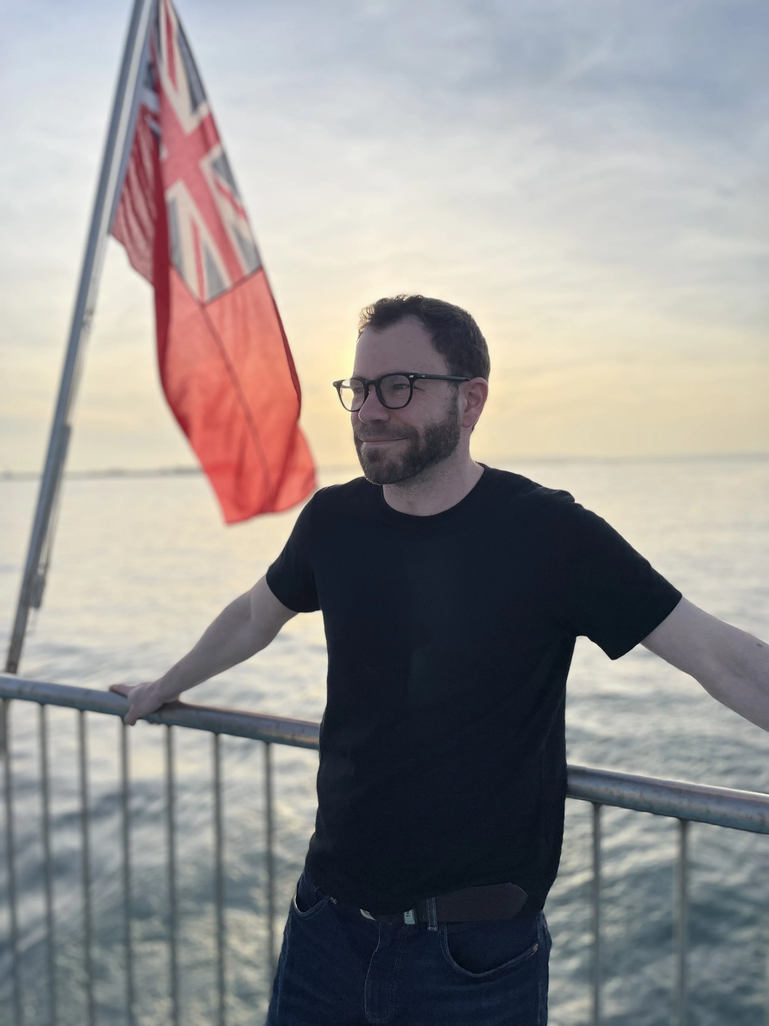 A man with glasses and a beard stands on a ship deck, smiling, with a British flag in the background during sunset over water.