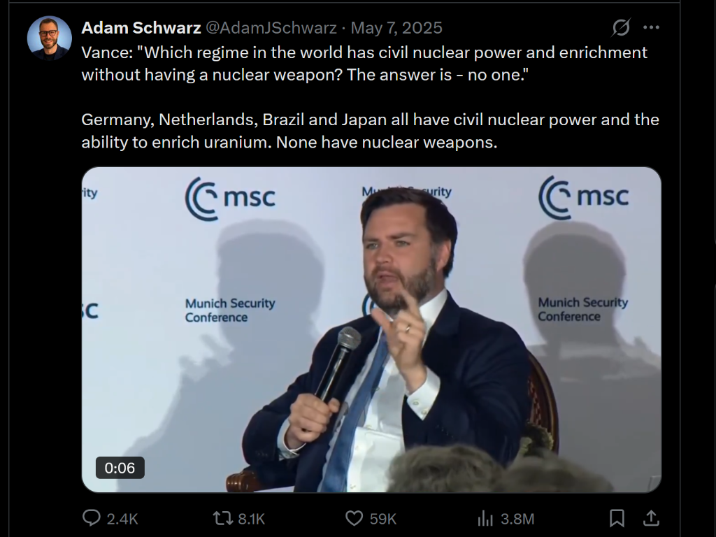 A man with dark hair and beard, wearing a dark suit and white shirt, speaking at a Munich Security Conference, holding a microphone, with a shadowed background featuring the Munich Security Conference logo.