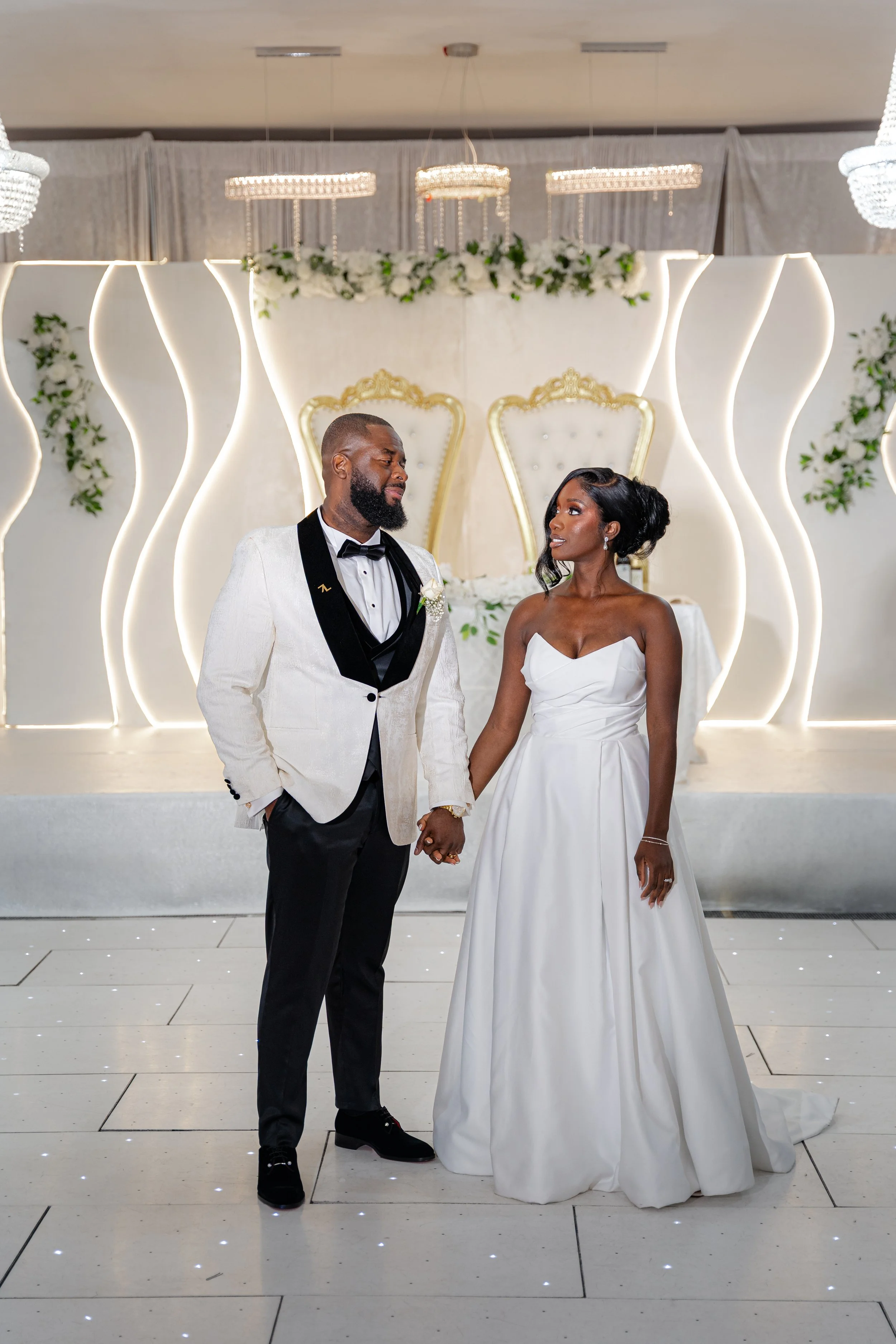 A bride and groom standing hand in hand during their wedding ceremony, exchanging vows, in front of an elegant decorated backdrop with two high-backed chairs and floral arrangements.