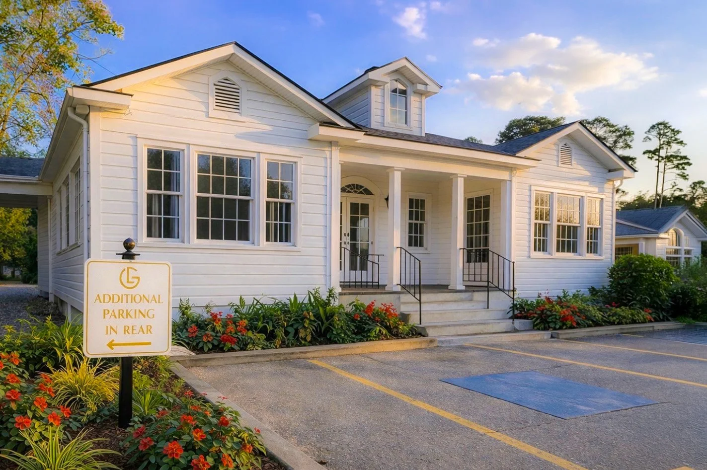White house with front porch and steps, surrounded by landscaping, with parking spaces and a sign indicating additional parking in rear.