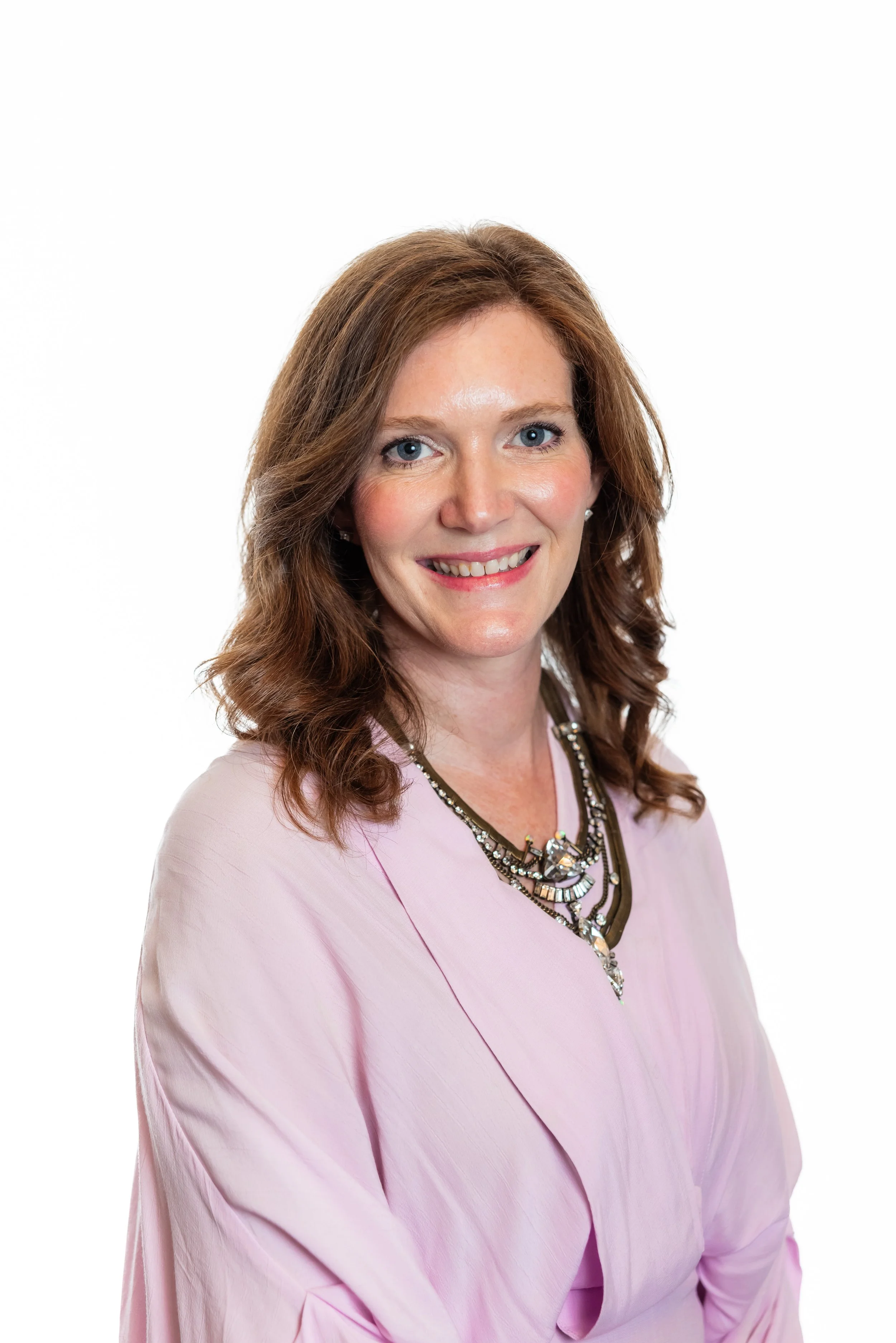 Portrait of a woman with light skin, wavy brown hair, blue eyes, and wearing a pink top and a statement necklace, smiling against a plain white background.
