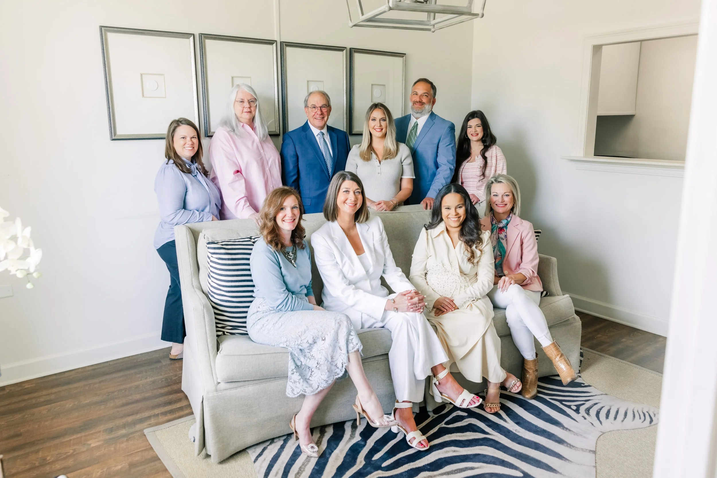 A group of ten diverse women and two men posing together in a living room, some sitting on a sofa and others standing behind, dressed in business casual attire, smiling for the photo.
