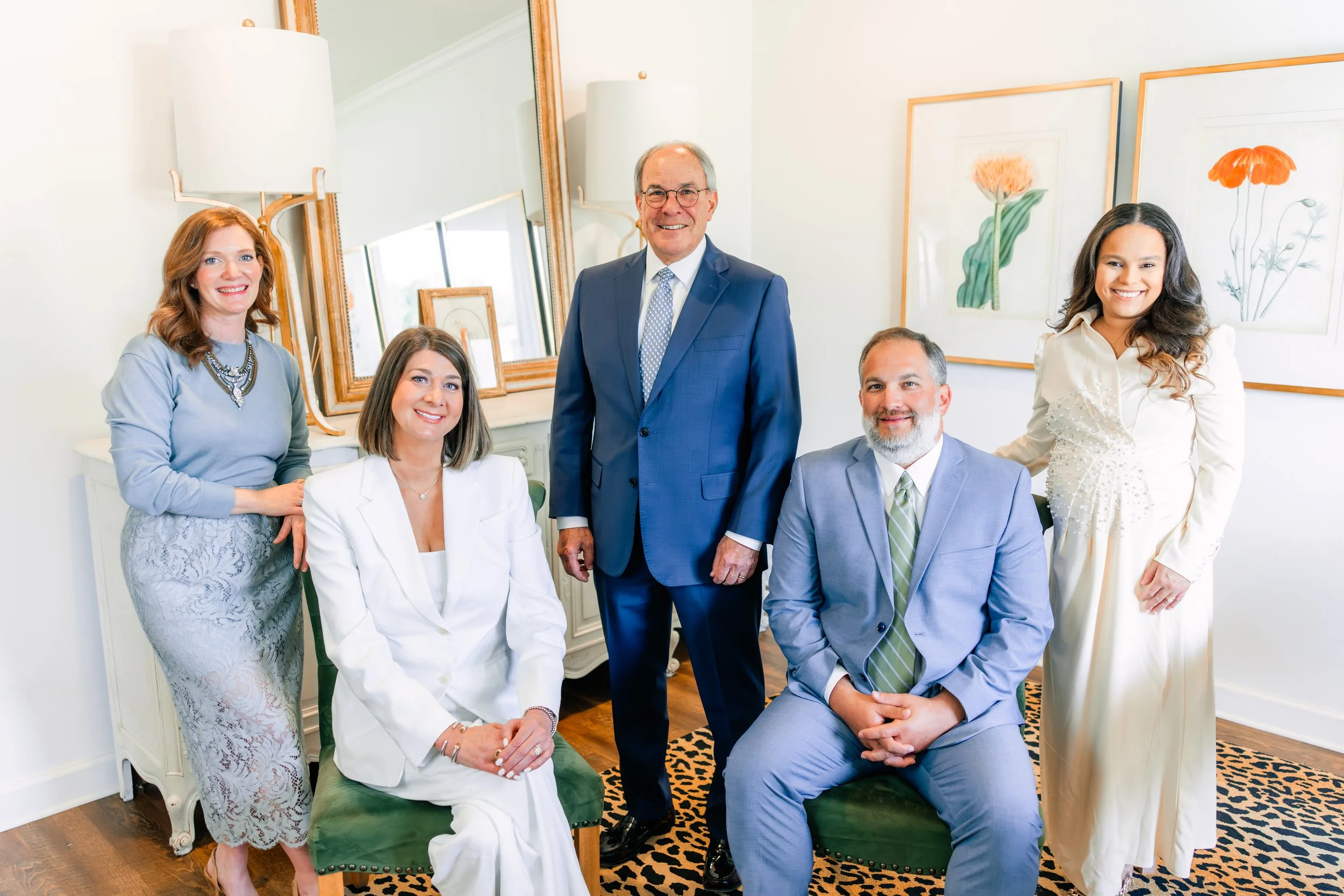 Group of six well-dressed people, three women and three men, posing indoors with framed botanical artwork on the wall behind them. One woman is seated on a green upholstered chair, while the others stand around her, smiling at the camera.