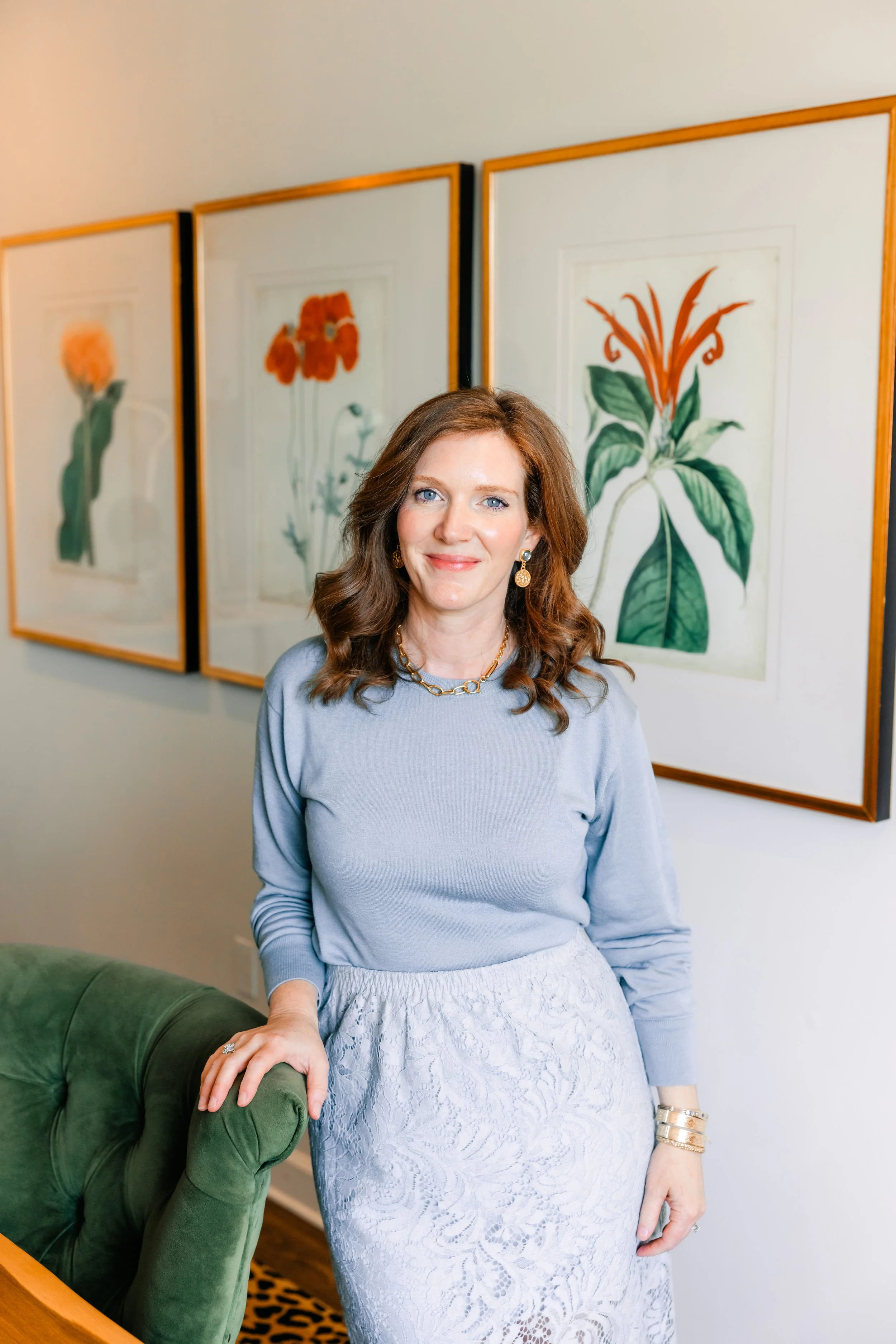 A woman with brown hair, wearing a light blue top, a white lace skirt, and gold jewelry, standing in an art gallery with botanical paintings in the background.